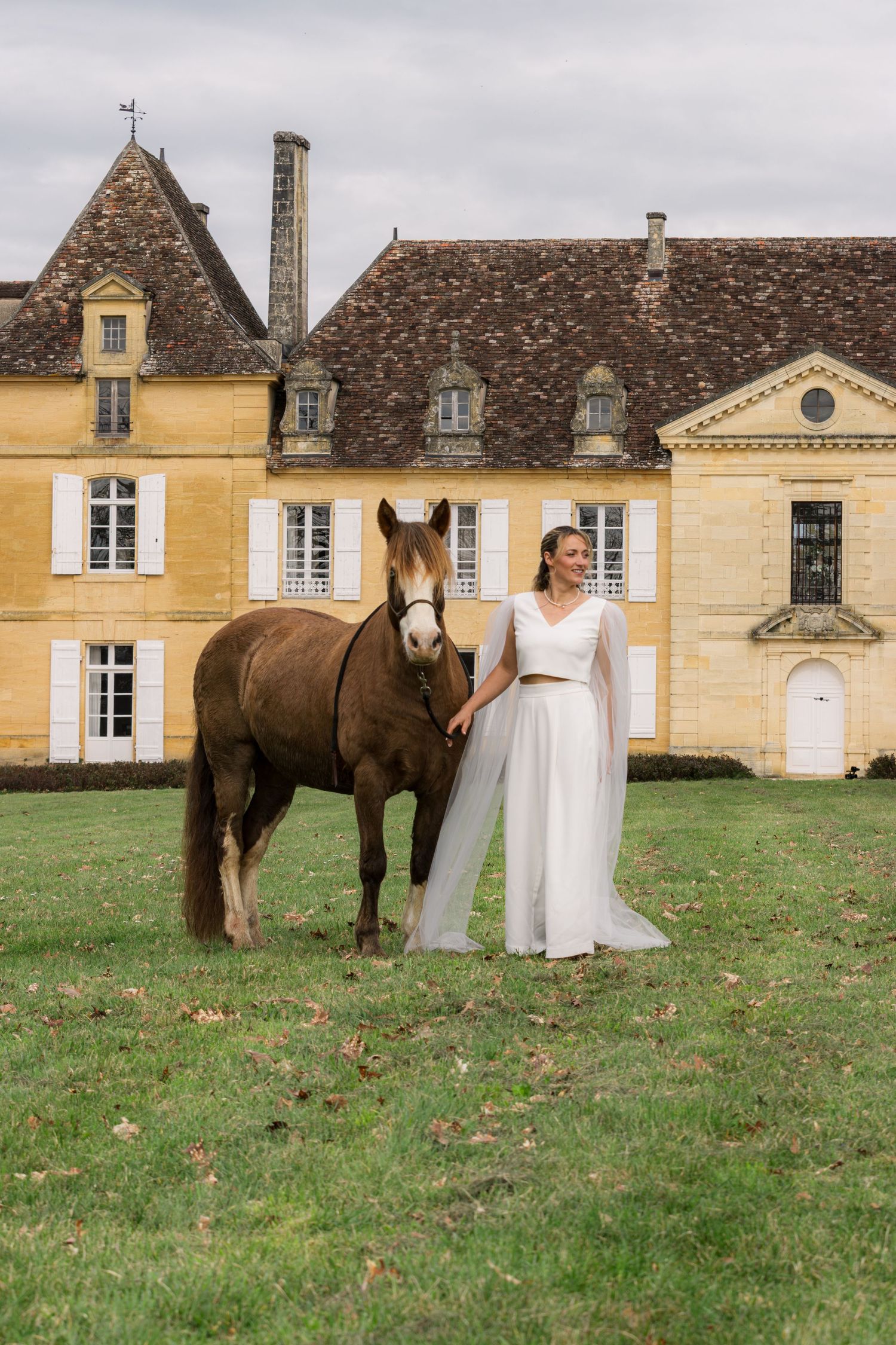 Bride in white gown stands beside a brown horse on green lawn before a grand yellow stone French chateau.