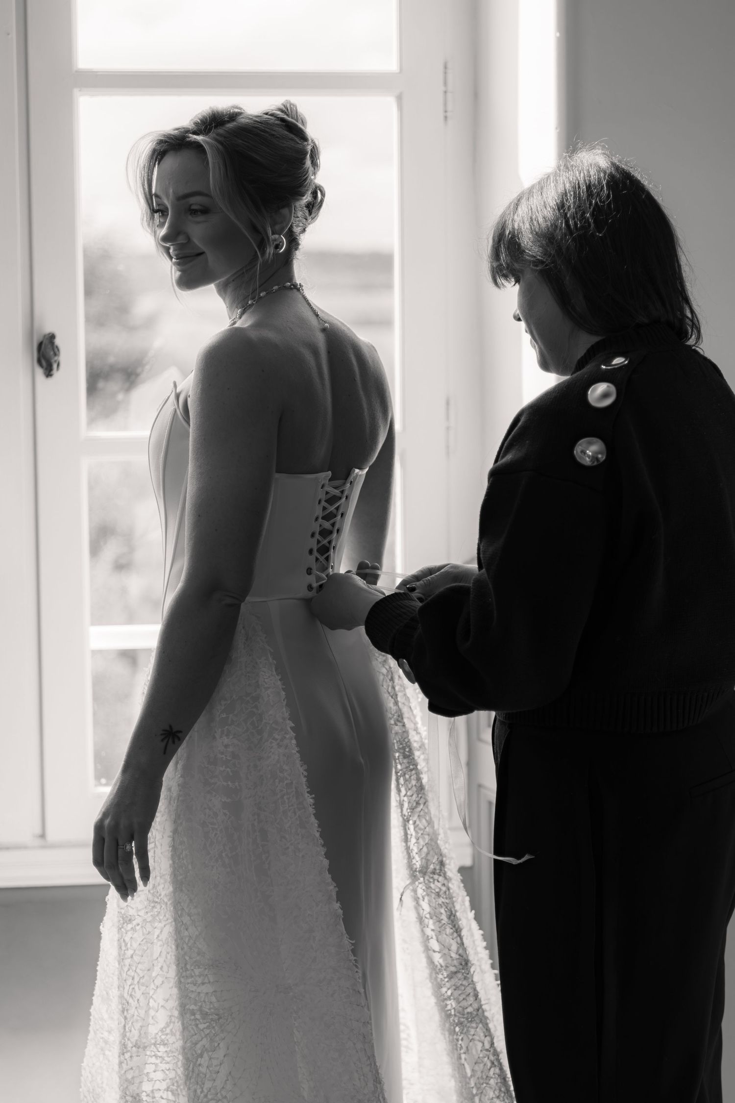 Black and white photo of a bride in a lace gown being helped with her dress by a woman near a bright window.