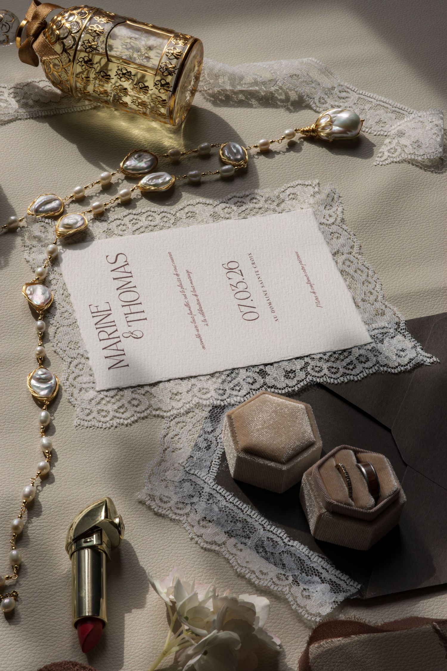 Elegant wedding invitation for Marine and Thomas dated 07.08.26, surrounded by gold jewelry, lace, lipstick, and ring boxes.