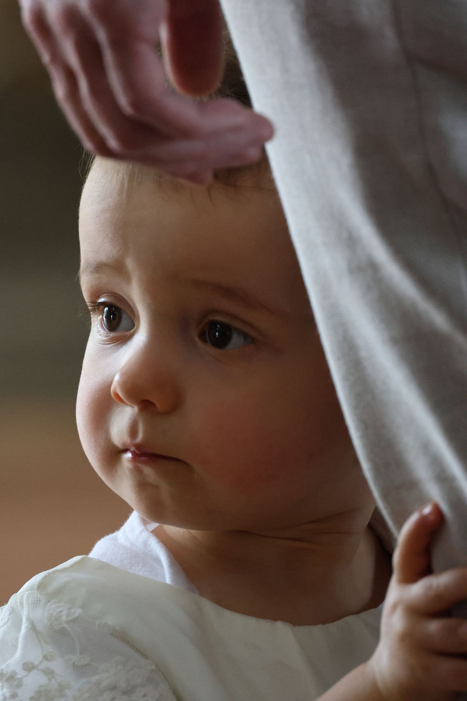 Curious baby with big brown eyes peeks around a white curtain, held gently by an adult hand.