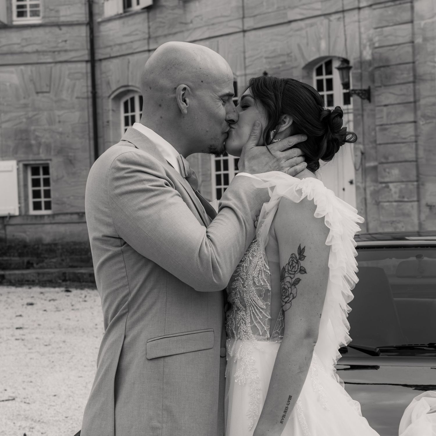 Black and white photo of a couple sharing a kiss at their wedding, bride in lace gown, groom in suit.