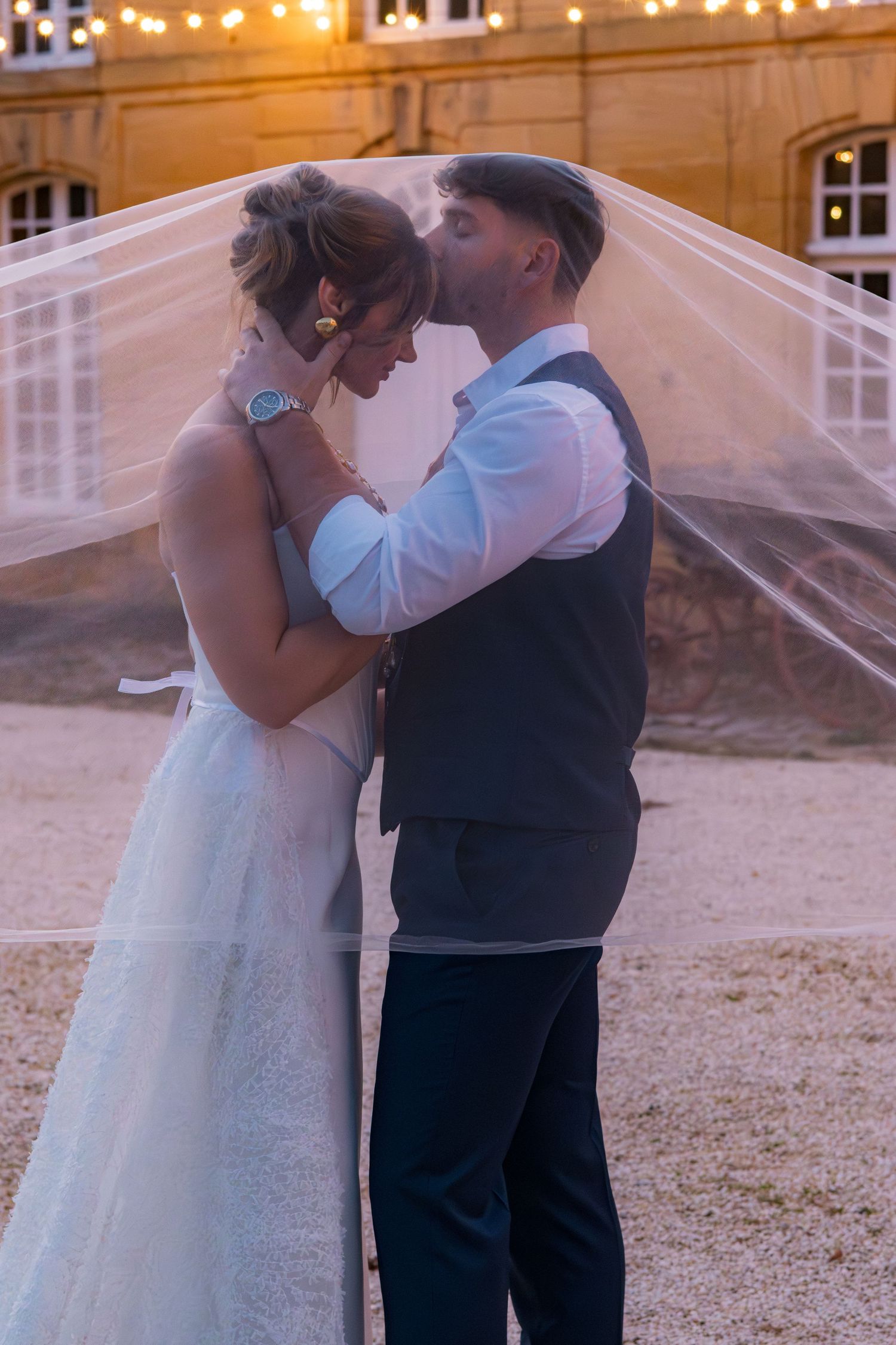 Bride and groom share a romantic moment under a flowing veil at dusk outside a warmly lit venue.