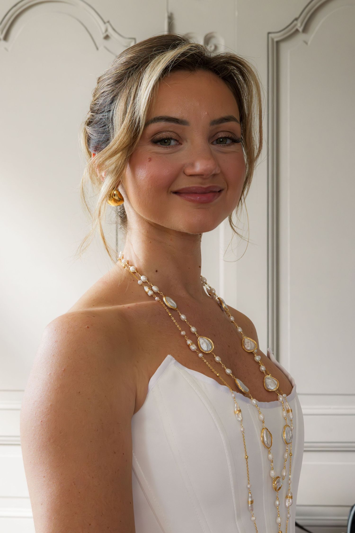 Elegant bride in white dress wearing layered pearl and gold necklaces with matching earrings, smiling softly.