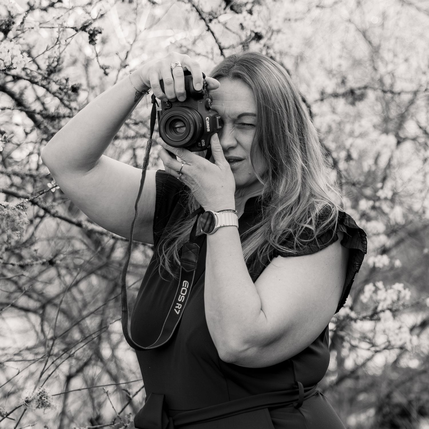Black and white photo of a woman holding a Canon camera up to her eye outdoors among blossoming trees.