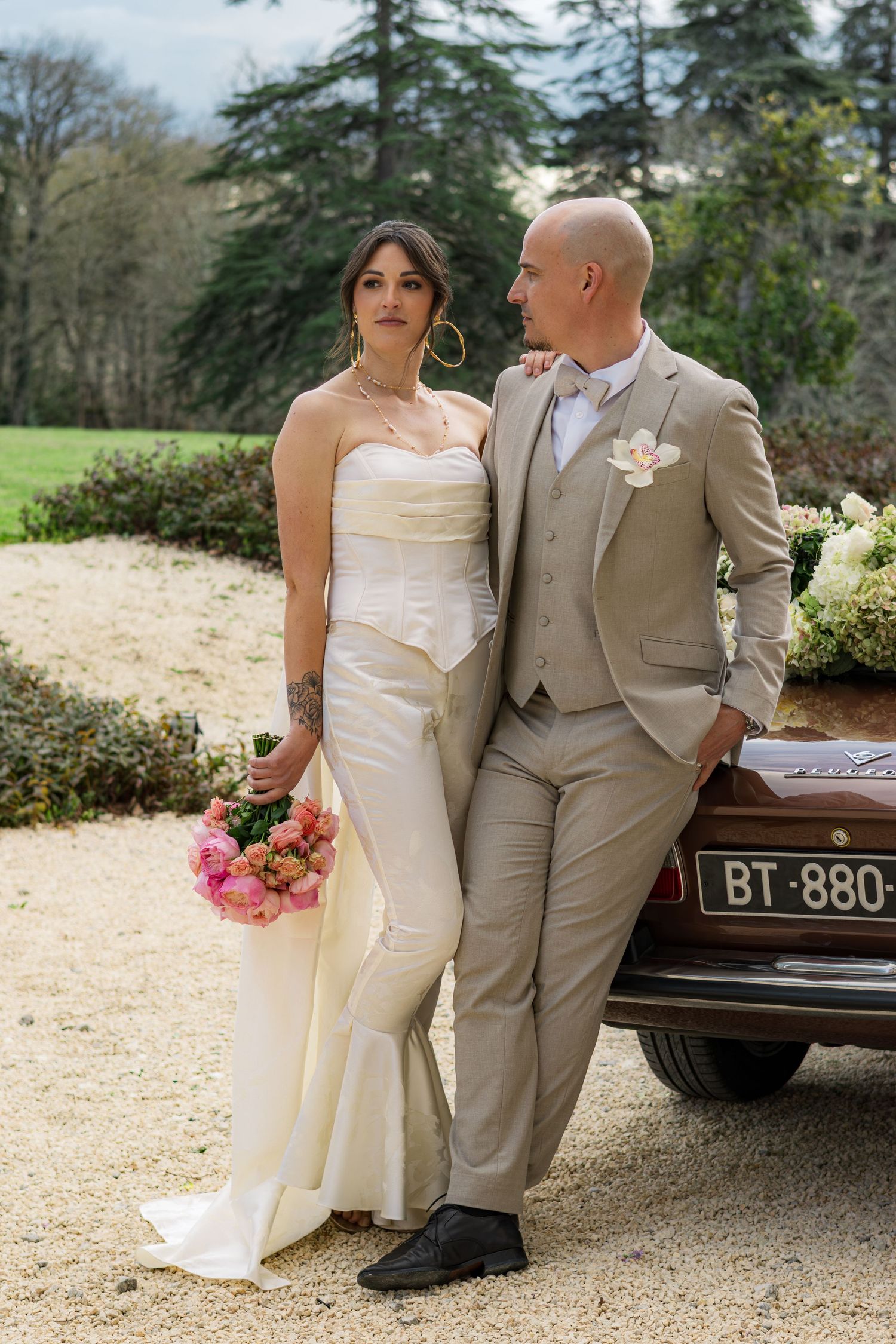 Bride in white gown holding pink bouquet poses with groom in tan suit beside vintage car in garden.