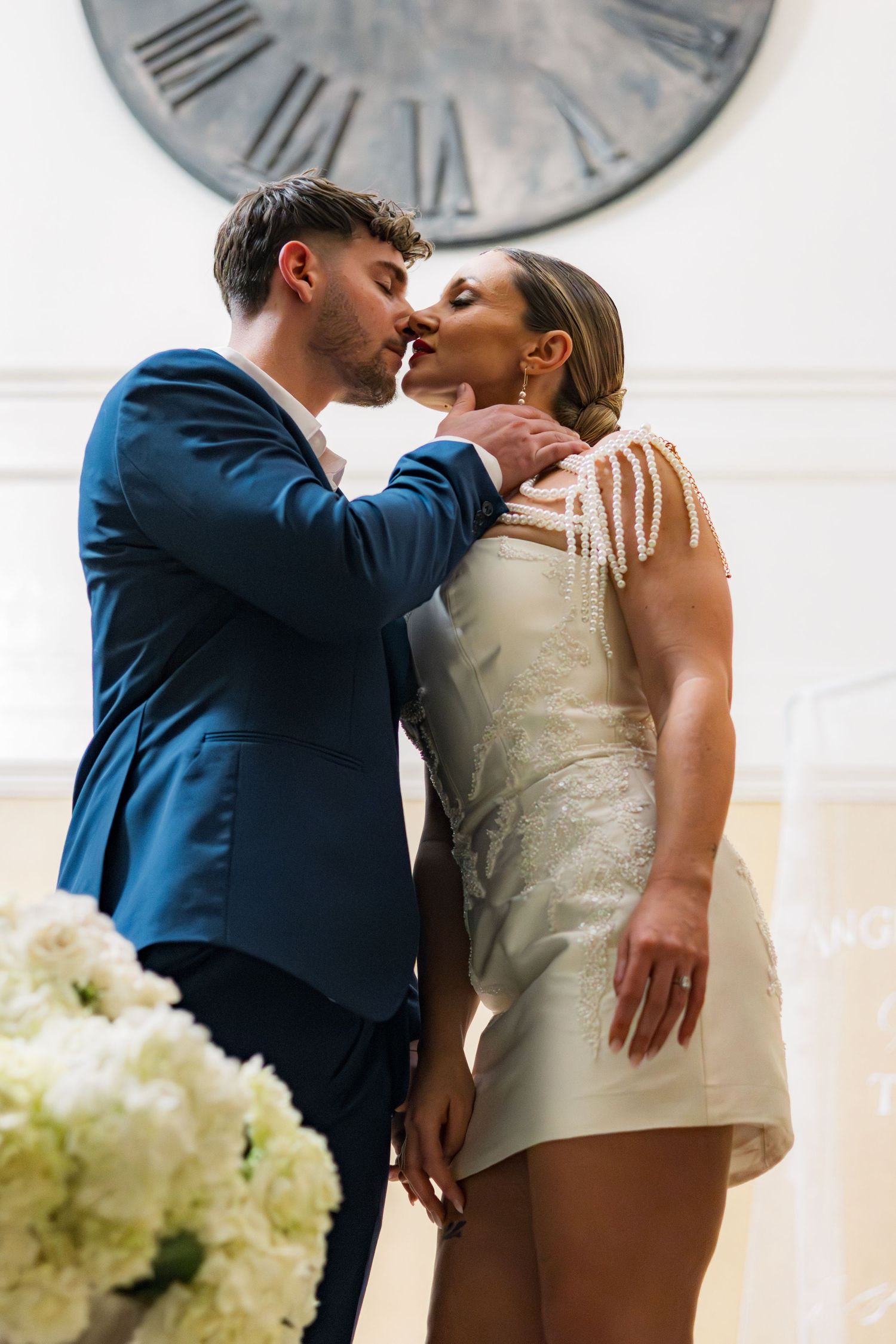 A couple shares a romantic kiss at their wedding, with the groom in a blue suit and bride in a short white beaded dress.