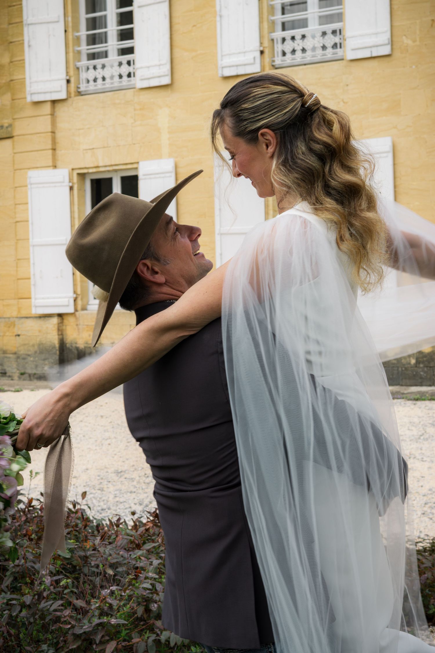 A groom in a wide-brim hat lifts his bride in a flowing veil outside a yellow French chateau.