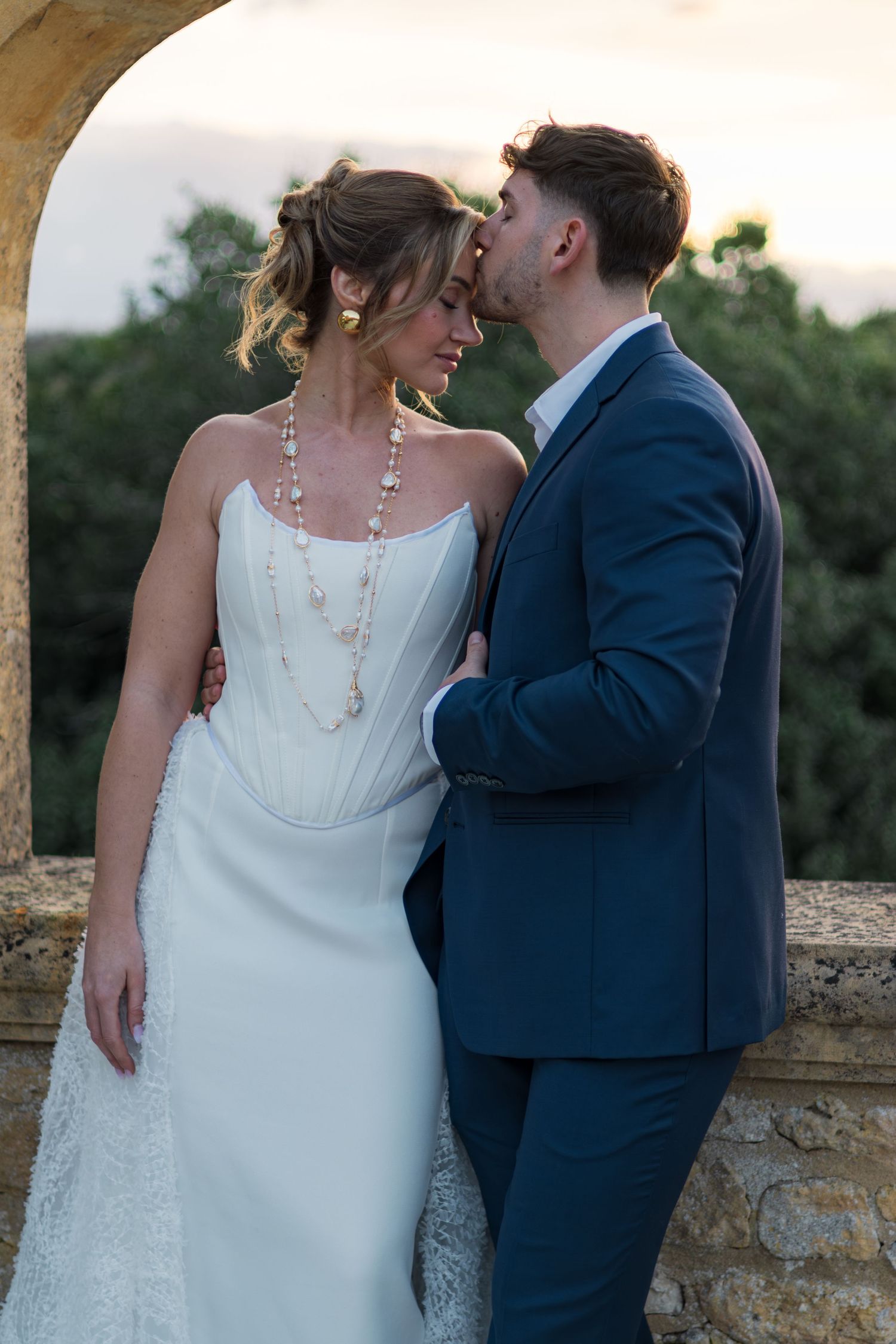 A bride and groom share a romantic moment outdoors, she in a white gown with pearls, he in a navy suit.