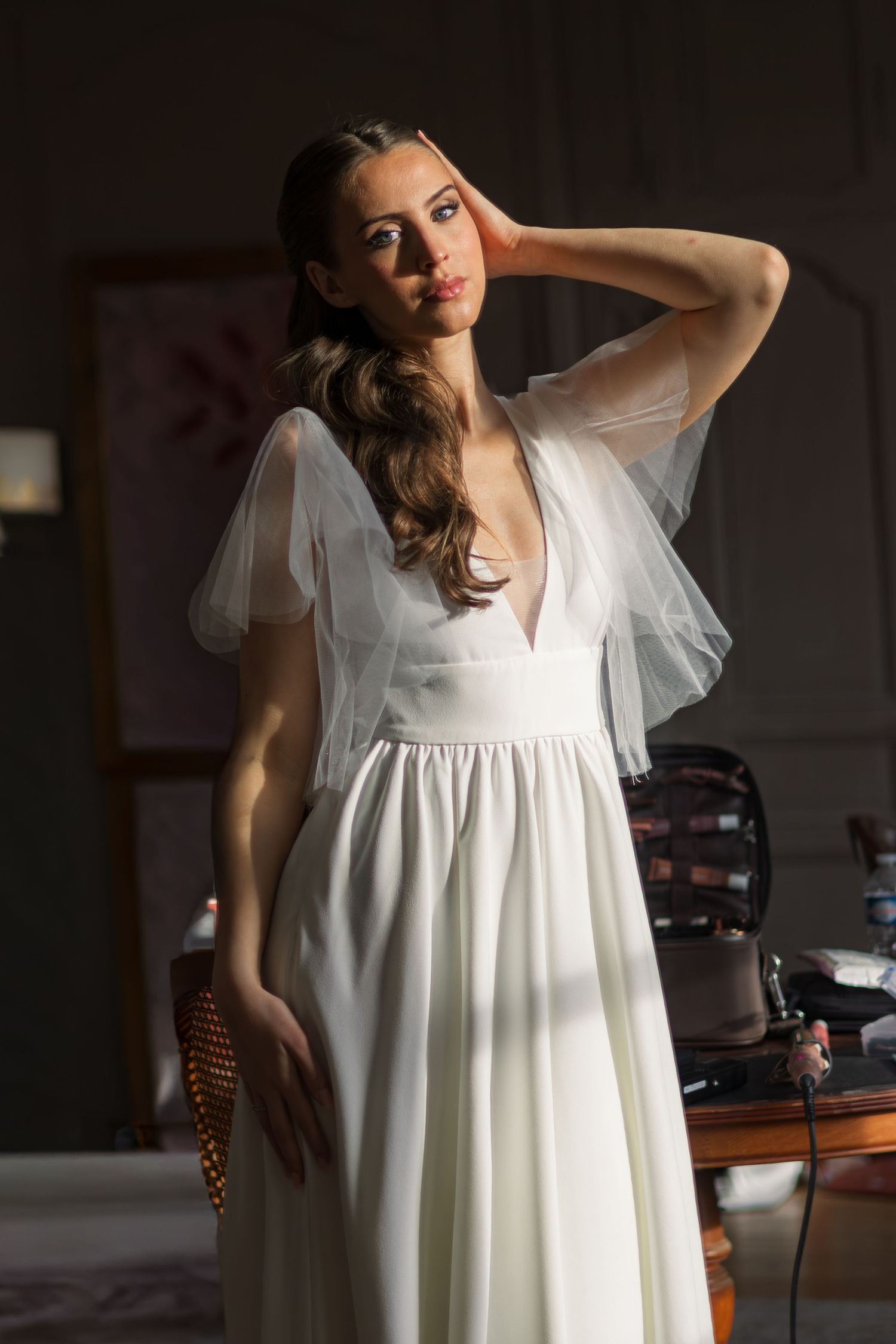 Elegant bride in a white chiffon gown with sheer sleeves, posing gracefully in a softly lit room.