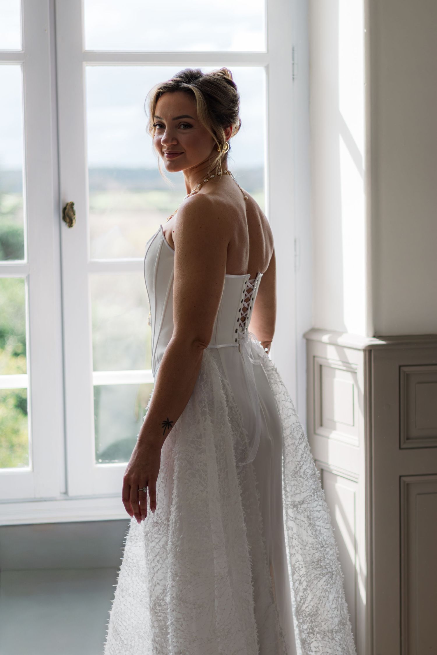 Bride in a white lace-back wedding gown stands by a sunlit window, smiling over her shoulder.