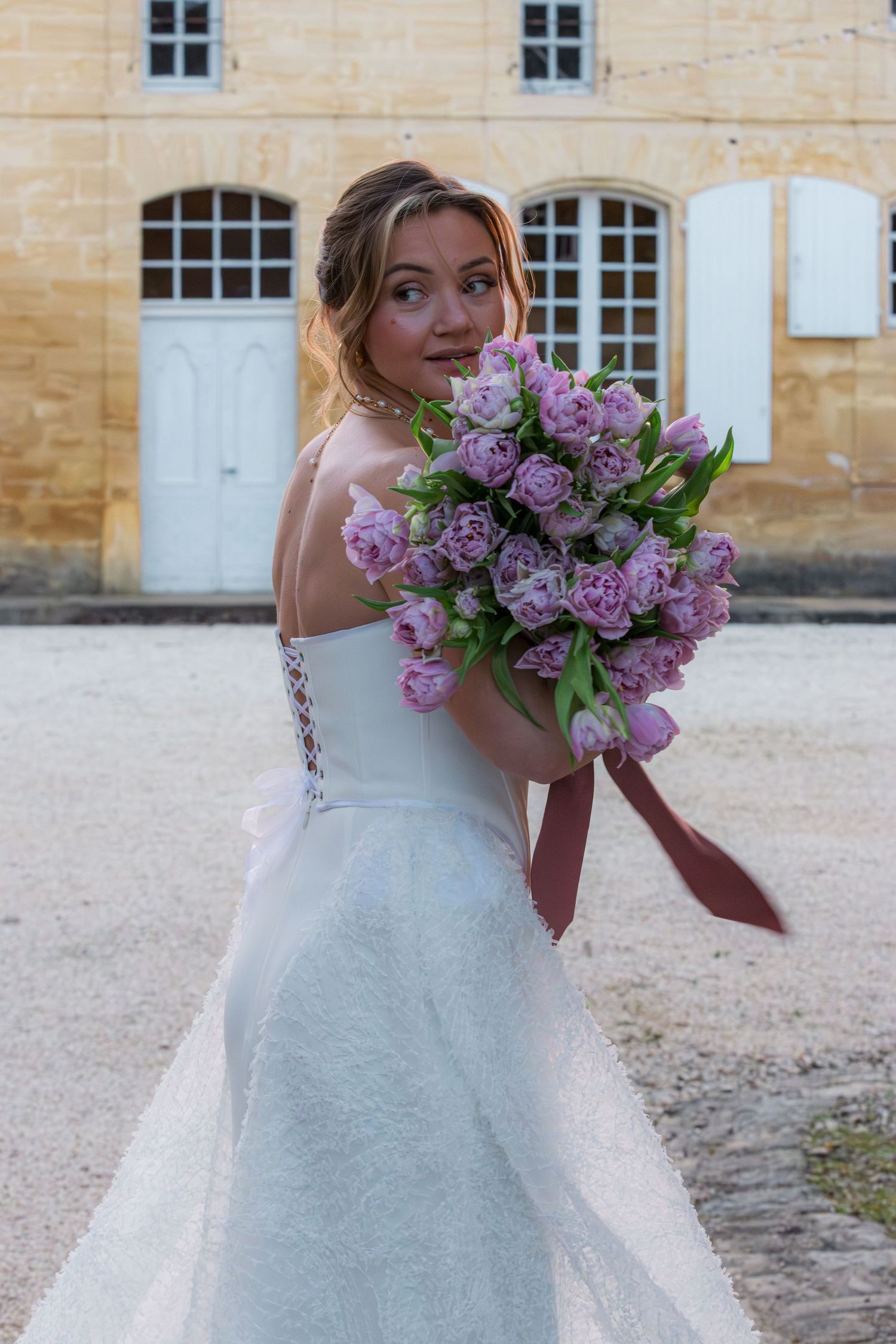 Bride in white backless gown holds purple tulip bouquet with pink ribbon, posing outdoors near a French chateau.