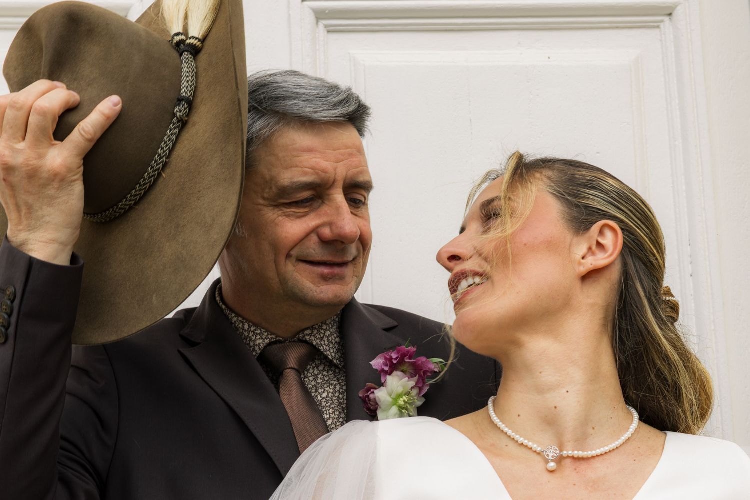 A groom lifts his brown fedora hat while smiling at his bride in a white dress and pearl necklace on their wedding day.