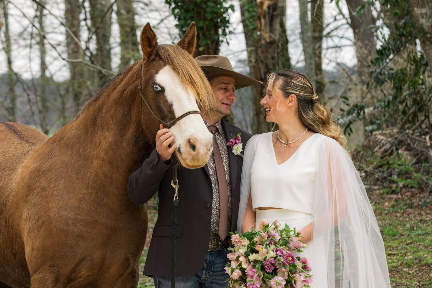 Bride and groom pose with a chestnut horse outdoors, bride holding a pink bouquet and wearing a veil.