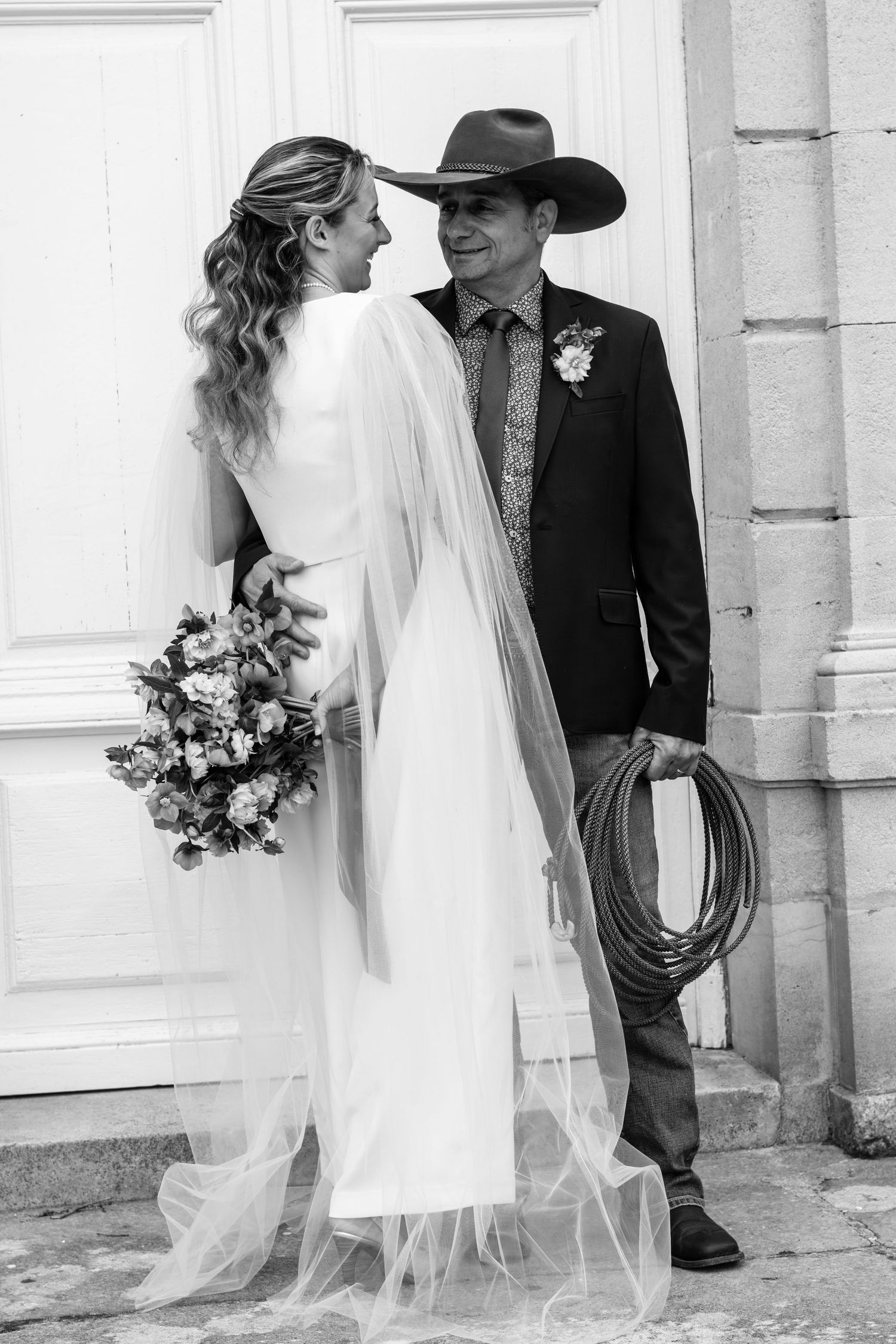 Black and white photo of a bride and groom, he wears a cowboy hat and holds a lasso, she holds a bouquet.