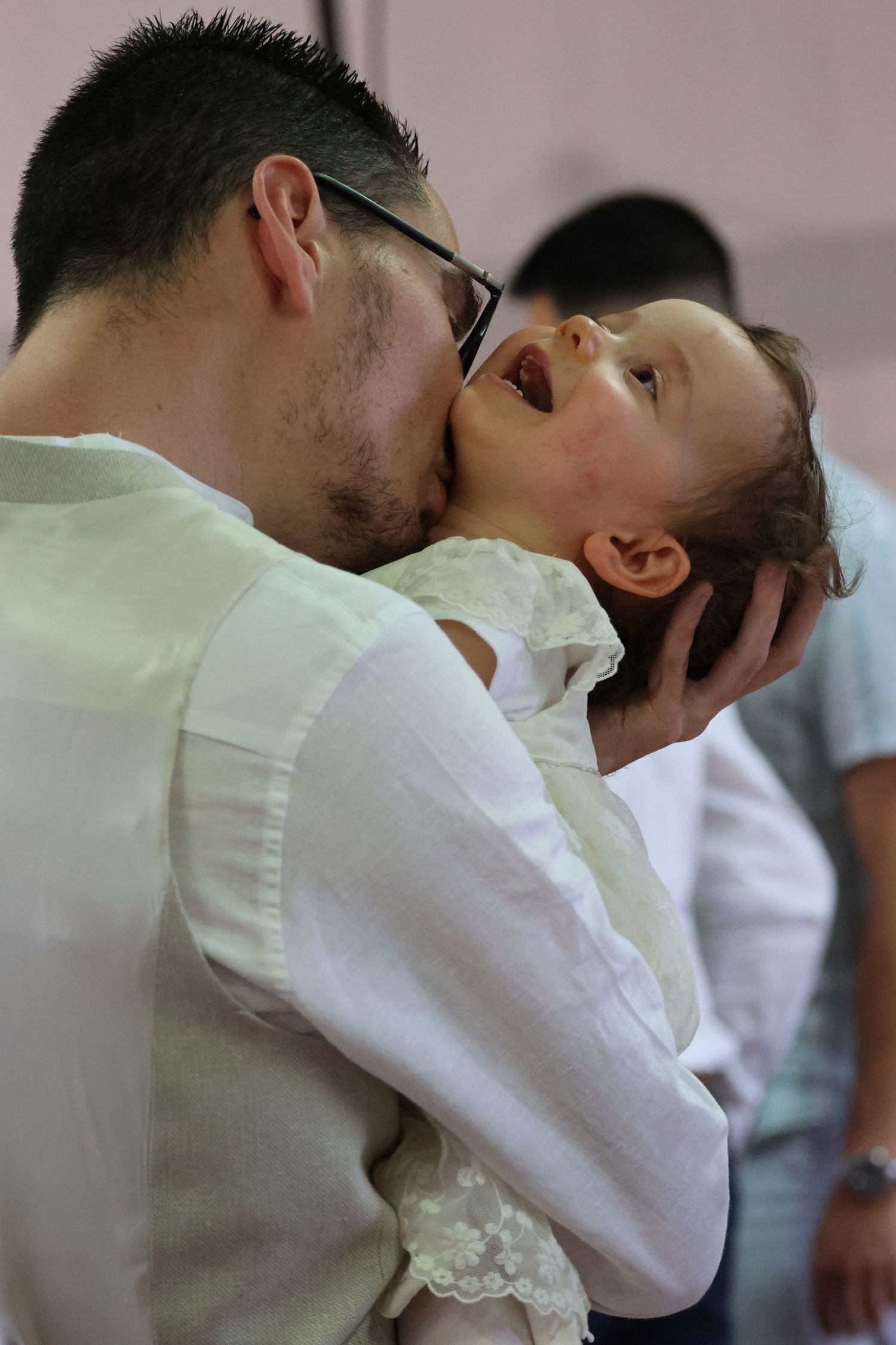 A man wearing glasses in a white shirt lovingly kisses a laughing baby he holds in his arms.