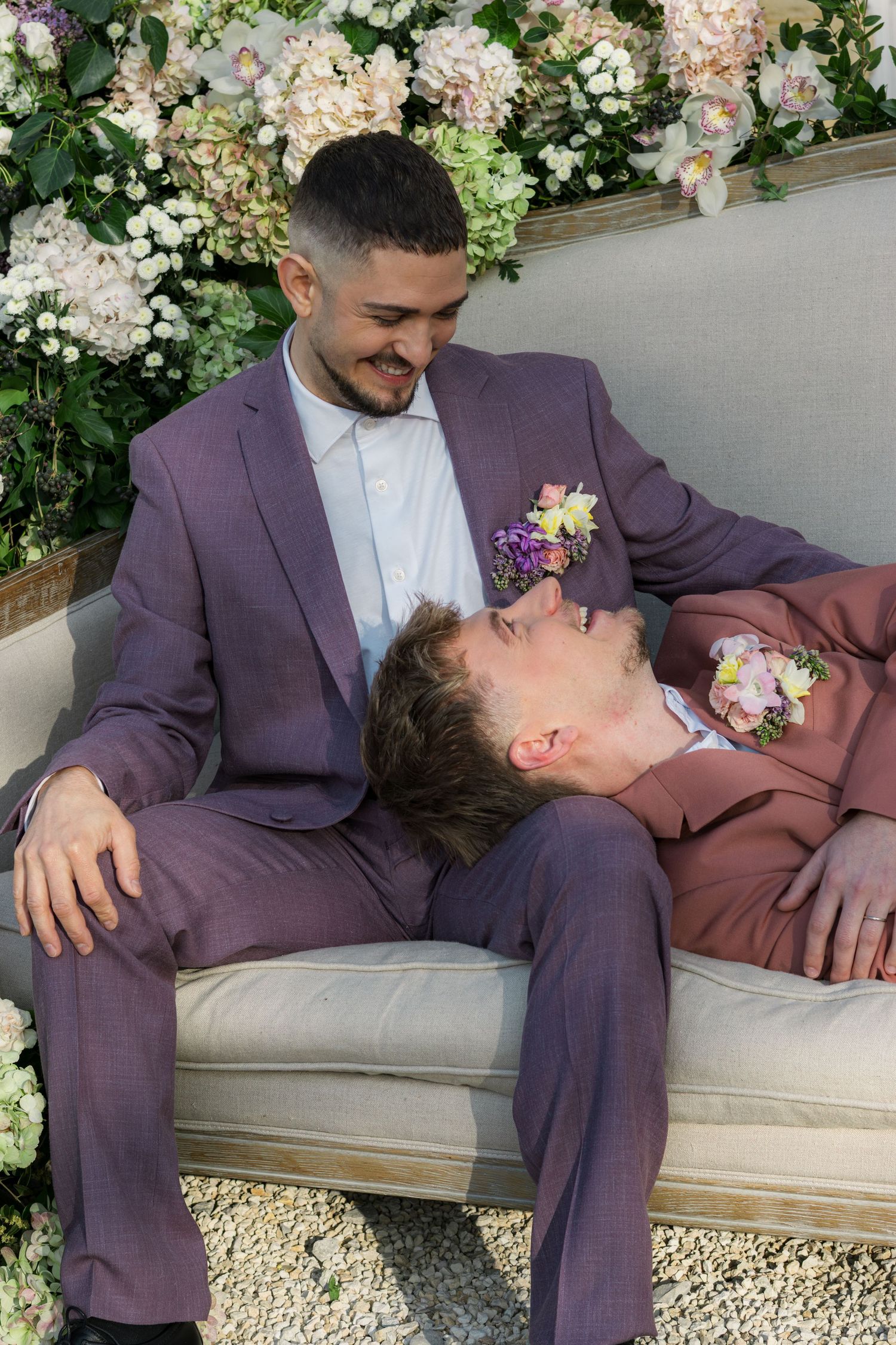 Two grooms in purple suits with floral boutonnieres sit on a vintage sofa surrounded by lush flowers.