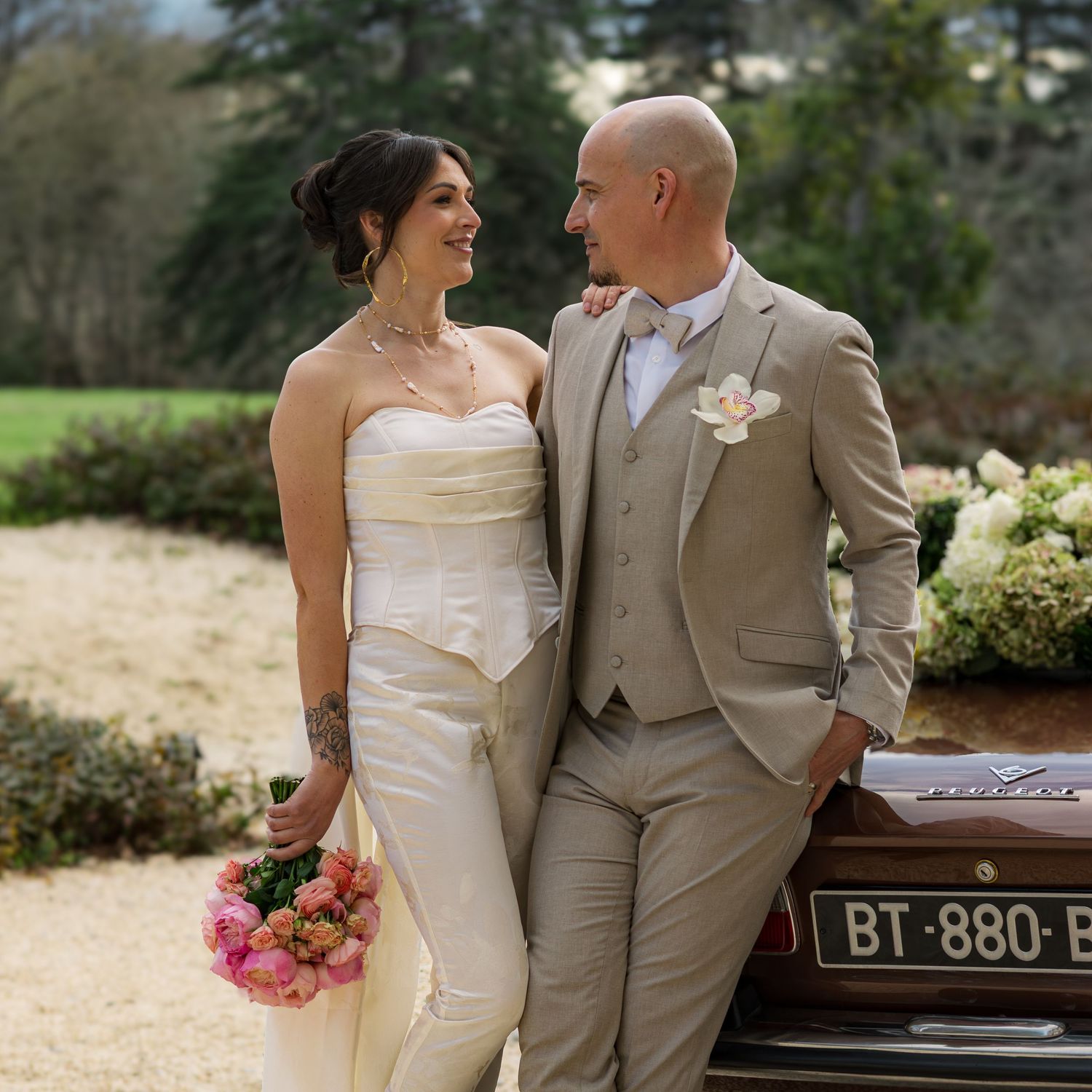 Bride and groom smiling at each other outdoors, she holds pink peonies in a white gown, he wears a tan suit.