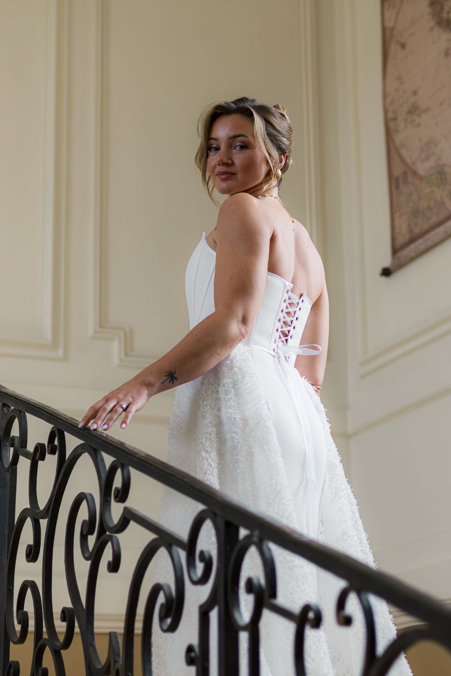 Bride in white lace corset gown descends elegant staircase with iron railing, glancing back gracefully.