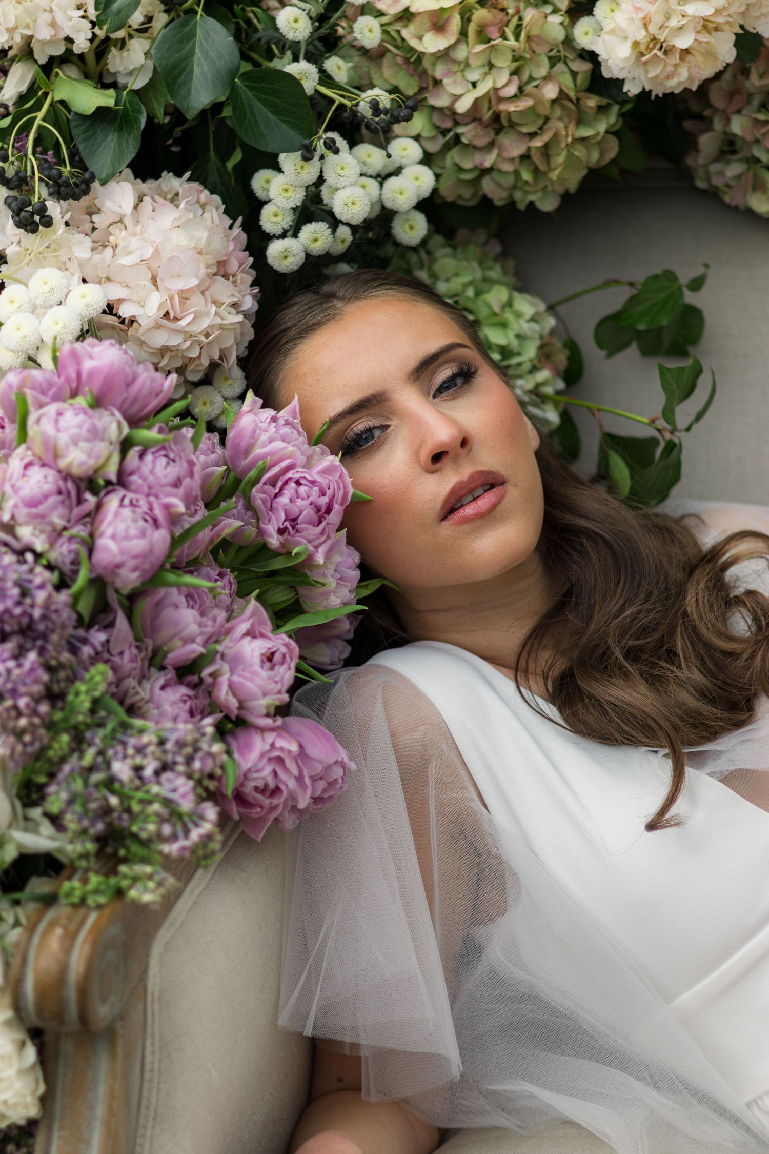 A bride in a white tulle dress reclines among lush purple peonies and hydrangeas, gazing upward dreamily.