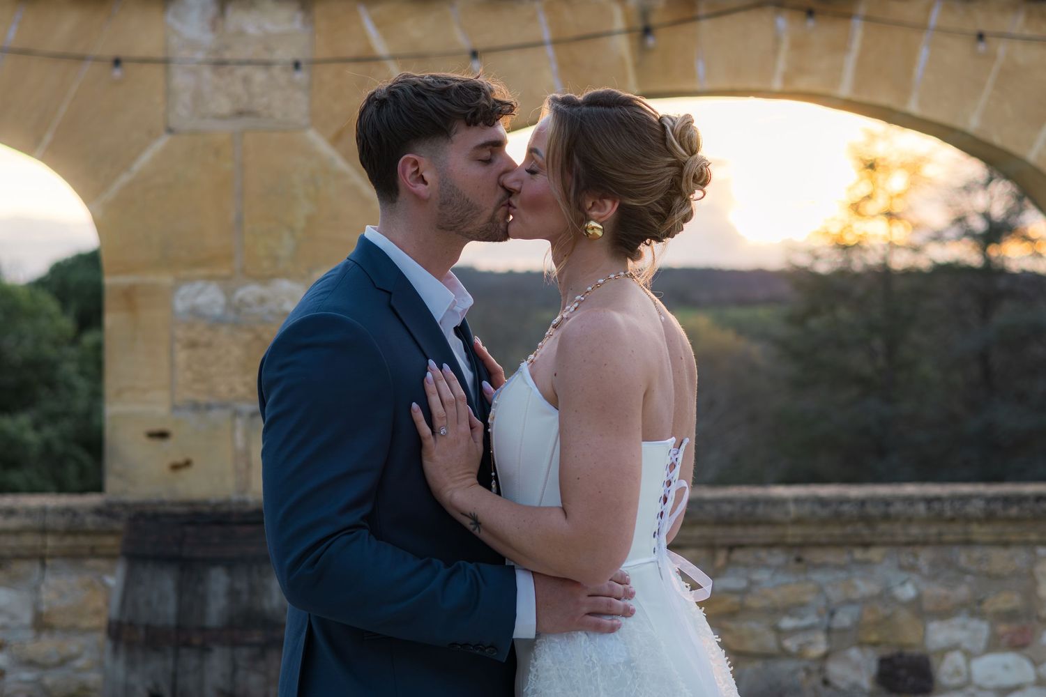 Bride and groom sharing a romantic kiss at sunset near a stone arch at their outdoor wedding ceremony.