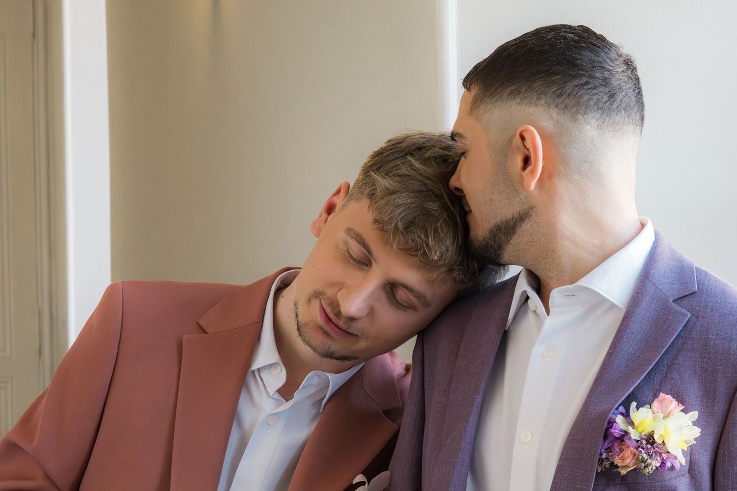 Two grooms in pink and purple suits share an intimate moment, one resting his head on the other's shoulder.