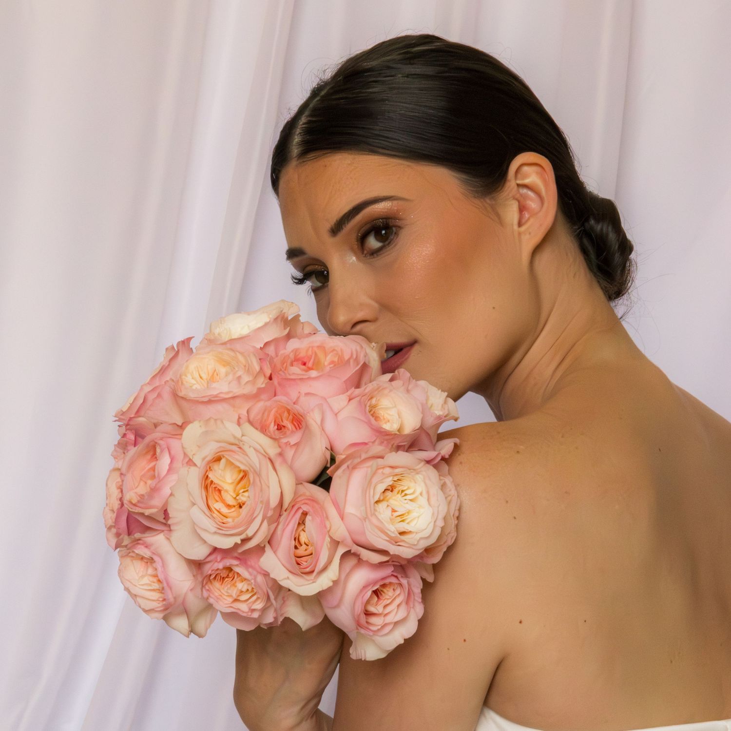 Elegant bride holding a lush pink rose bouquet against a white backdrop, showcasing natural bridal beauty.