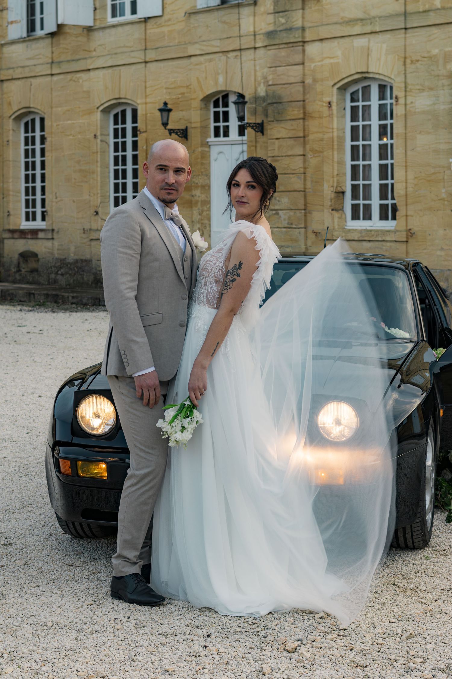Bride and groom pose beside a classic black sports car in front of an elegant French chateau on their wedding day.
