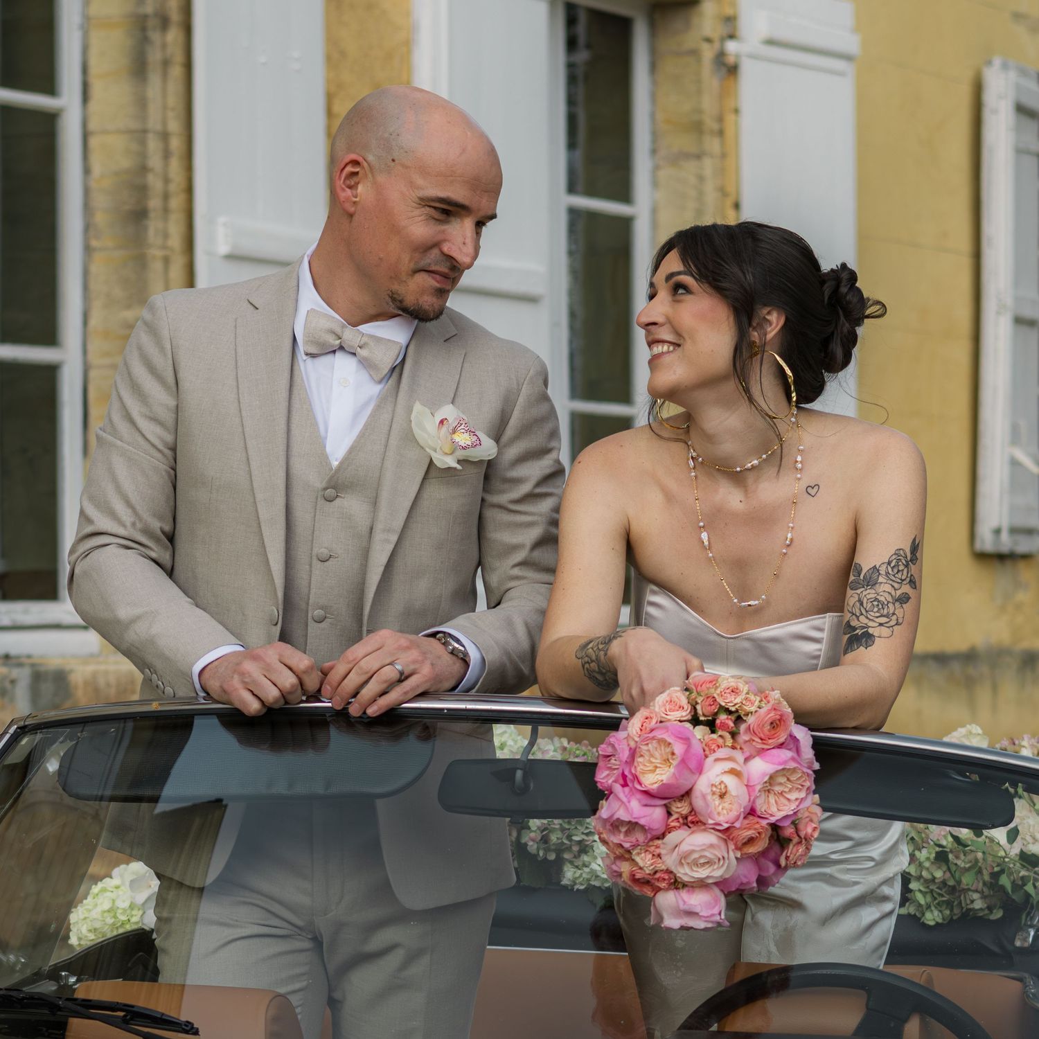 A bride and groom in wedding attire smile at each other beside a vintage car, bride holding a pink bouquet.