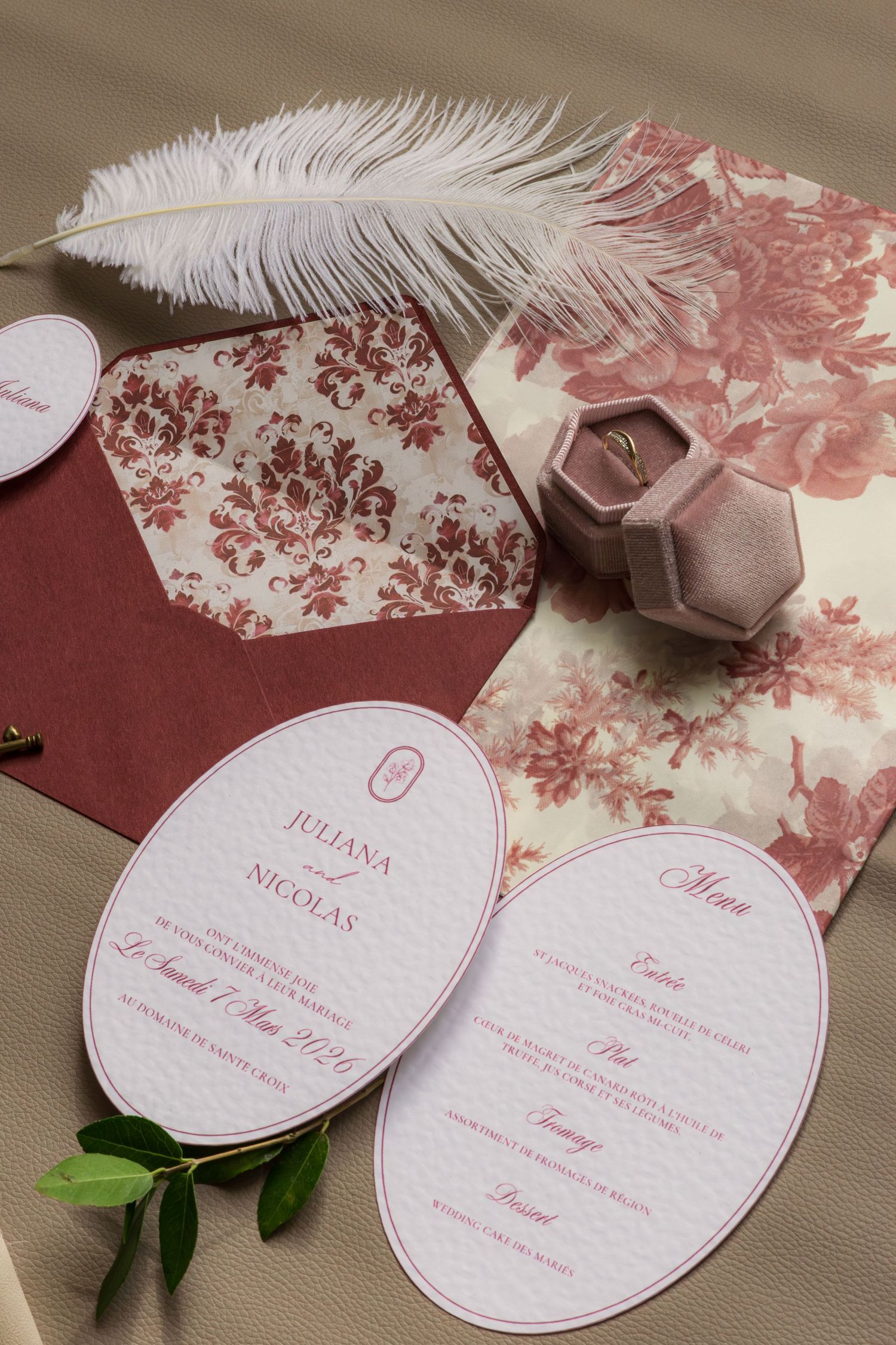 Elegant wedding stationery set with burgundy floral envelope, oval invitation cards, menu, and velvet ring box on beige background.