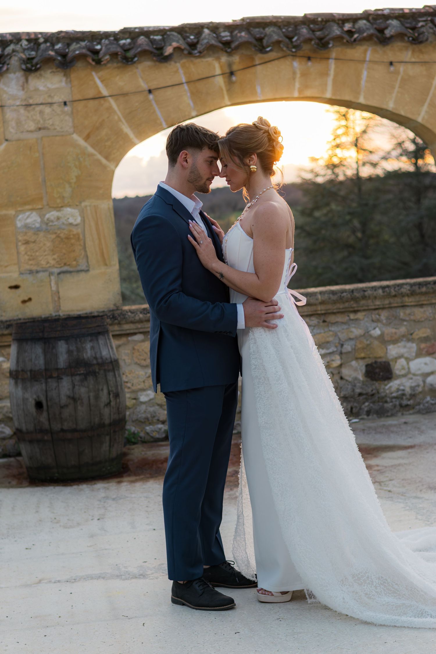 Bride and groom share a romantic sunset moment in front of a stone arch at an outdoor wedding venue.