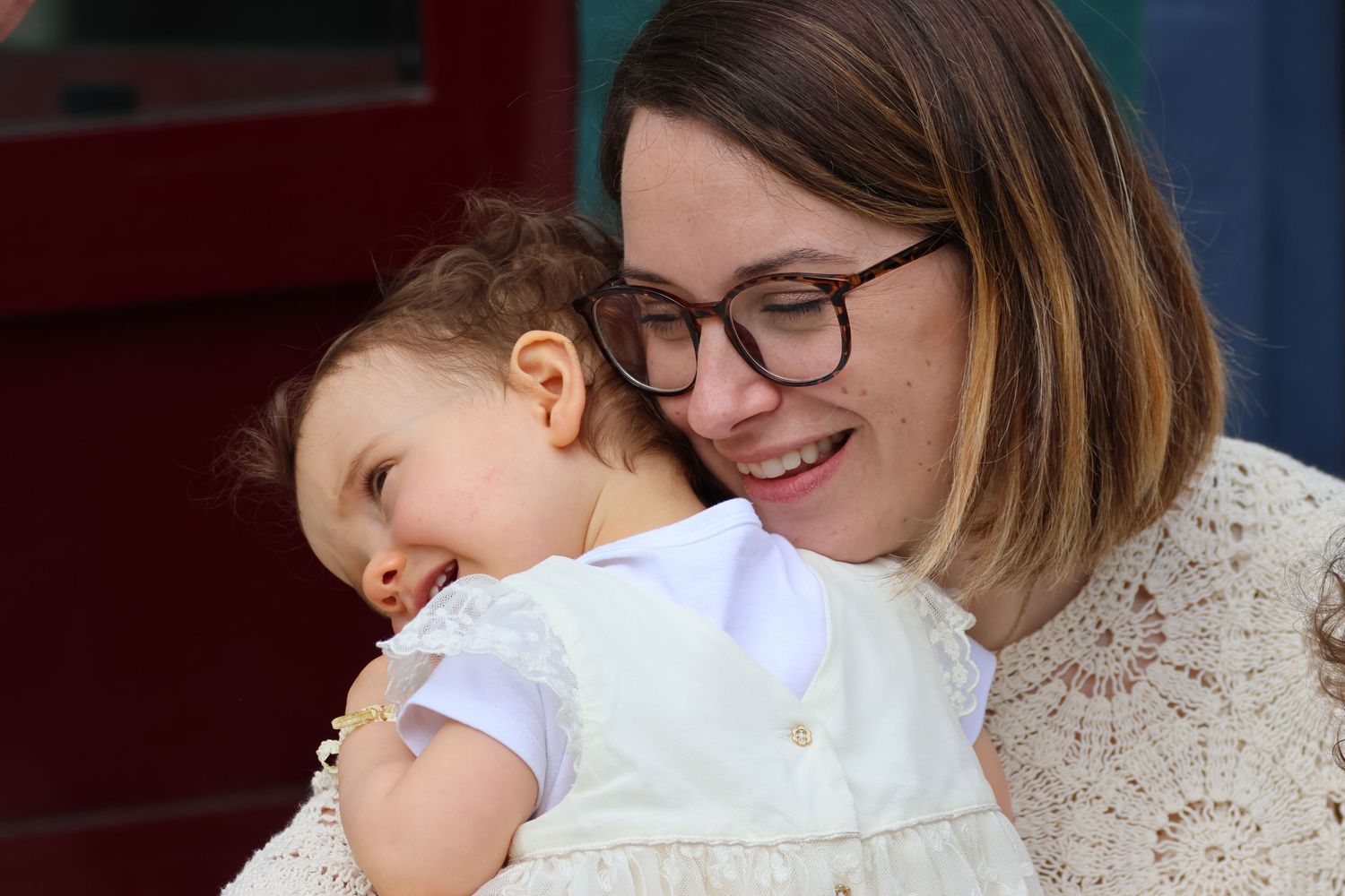 Woman with glasses smiling while holding a sleeping baby dressed in white.