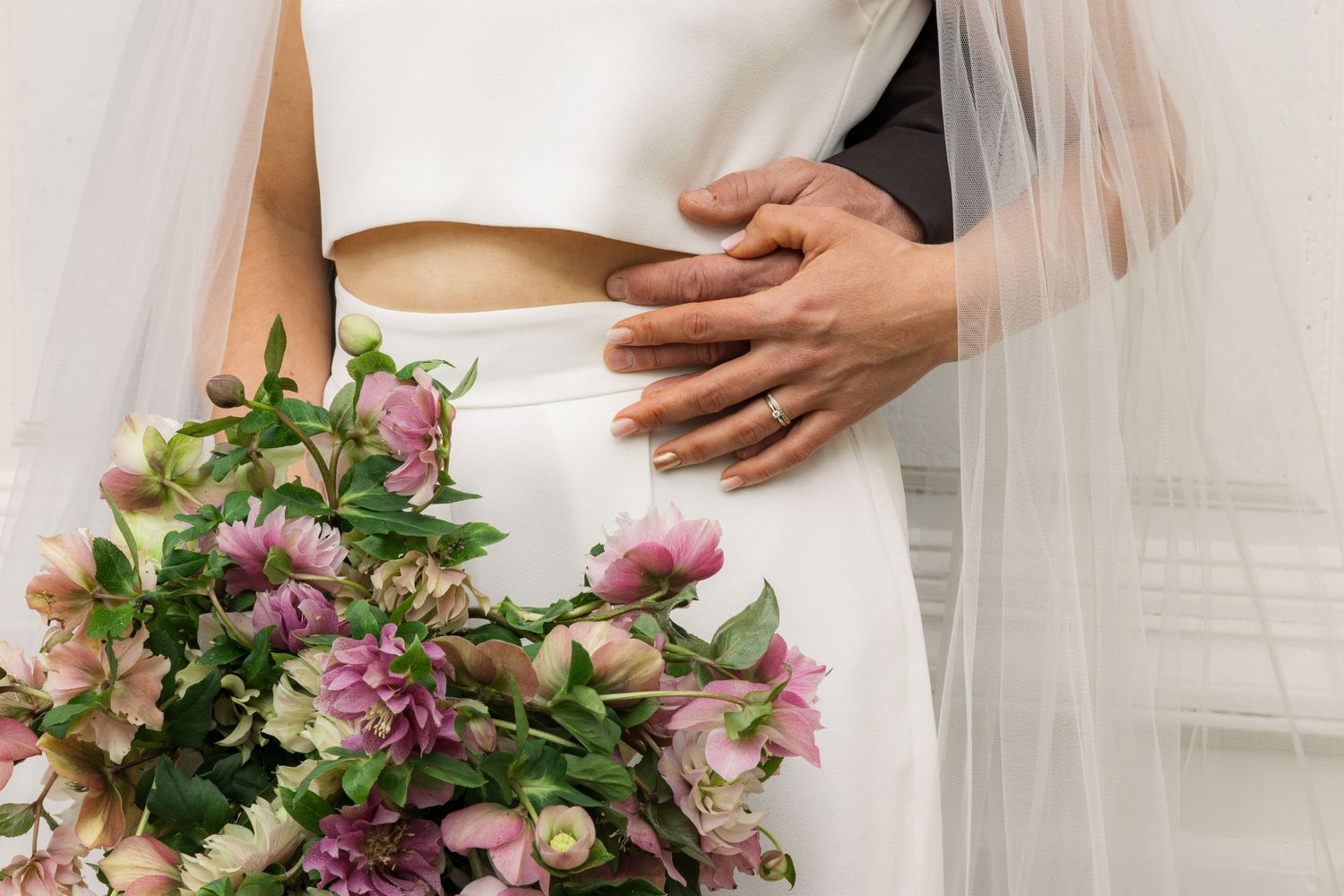 Bride in white two-piece gown holding pink floral bouquet with groom's hand on her waist at wedding.