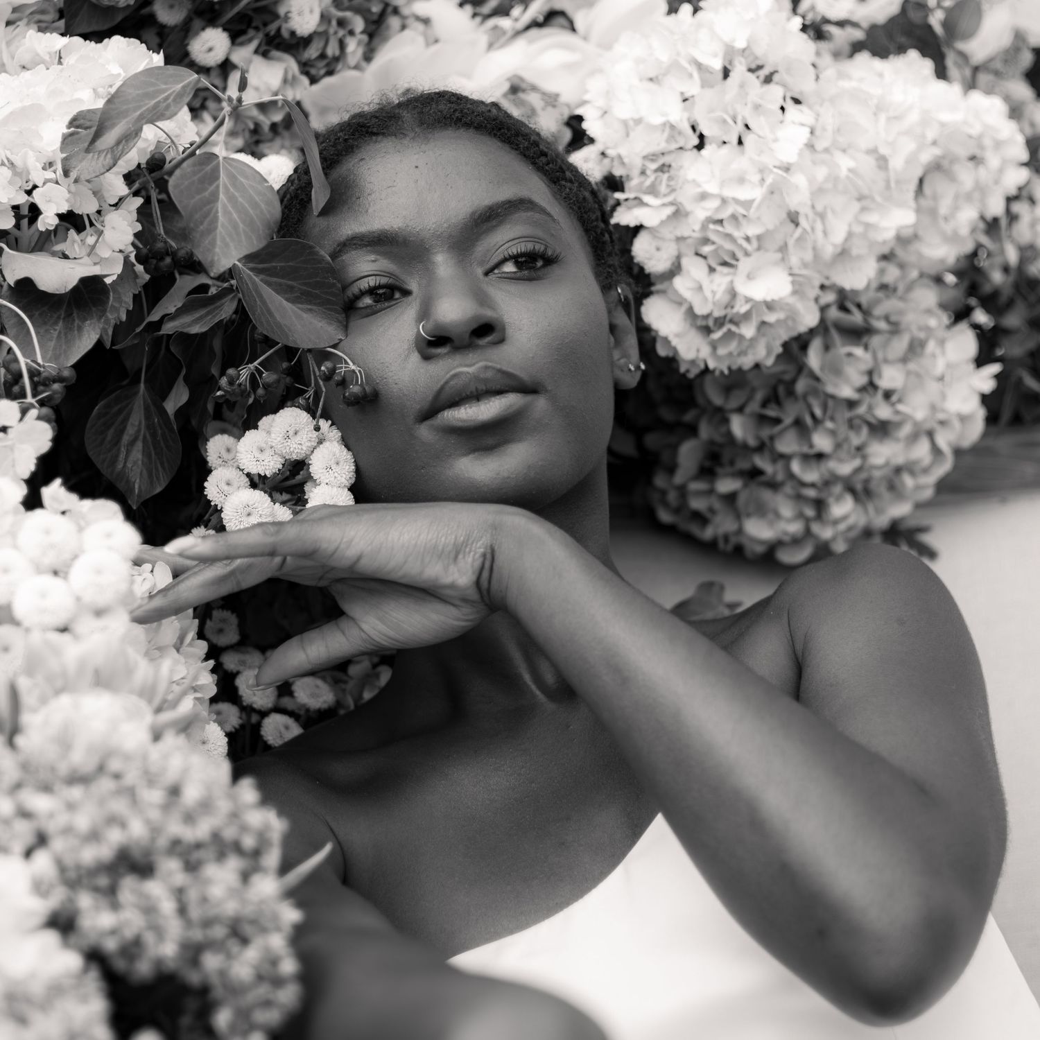Black and white portrait of a woman surrounded by lush hydrangea flowers, resting her chin on her hands.