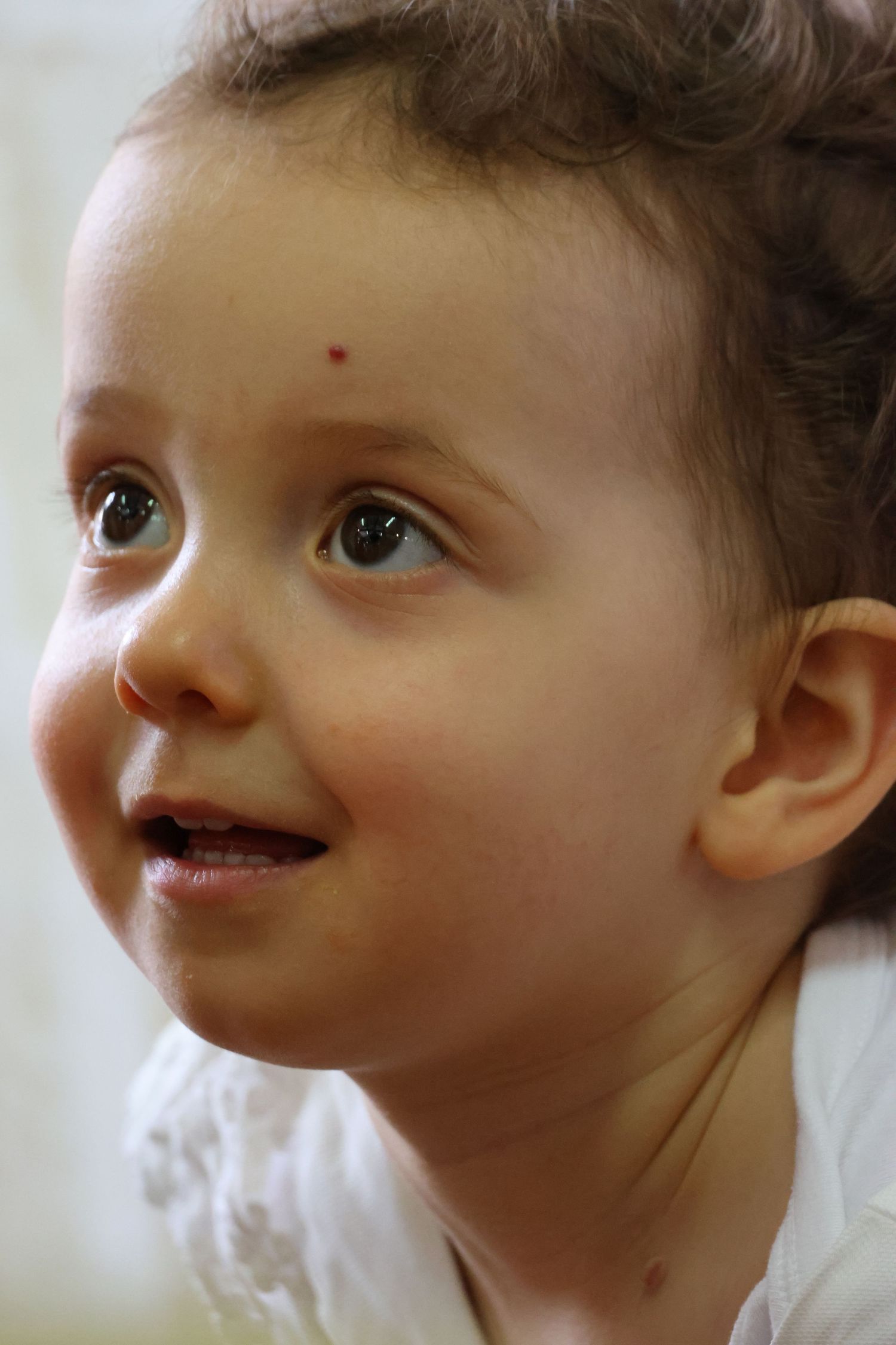 A curious toddler girl with brown eyes and a small red mark on her forehead smiles gently while looking upward.