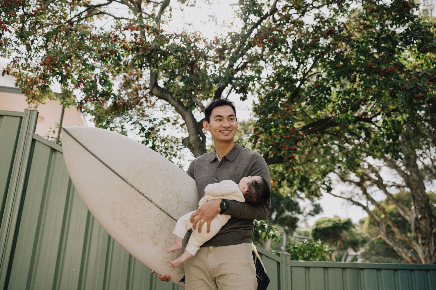 Smiling father holding a baby and surfboard outdoors near a green fence with lush trees in the background Sunshine Melbourne.