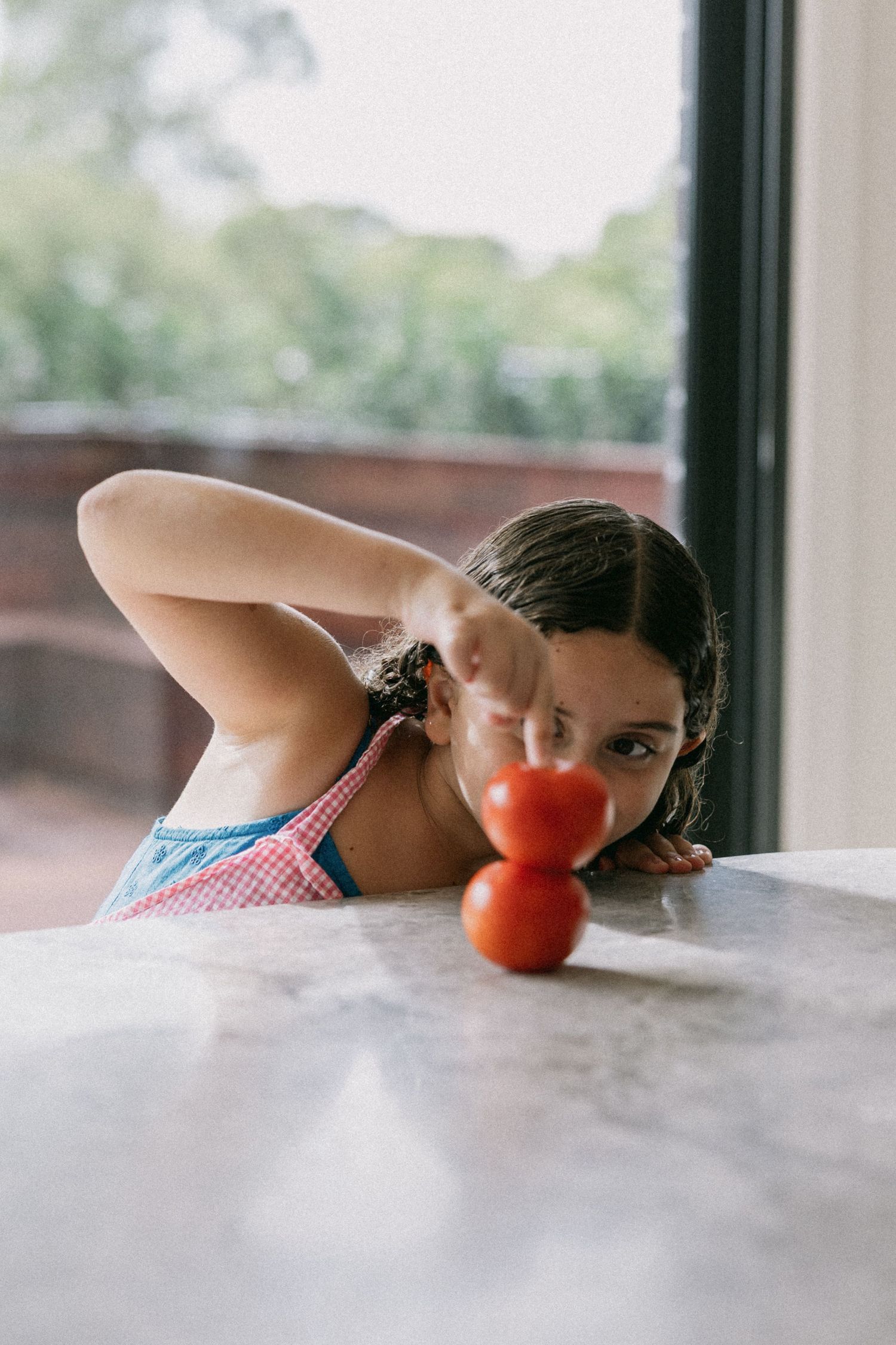 Young girl peering over a marble countertop at two red tomatoes near a window.