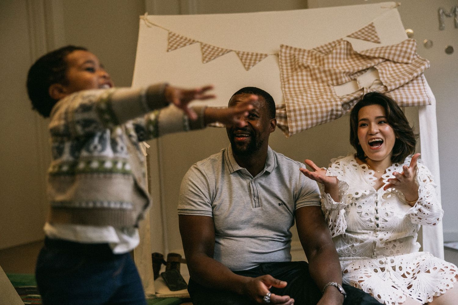 A joyful family moment with parents laughing as their young child plays and reaches out in a cozy decorated room.