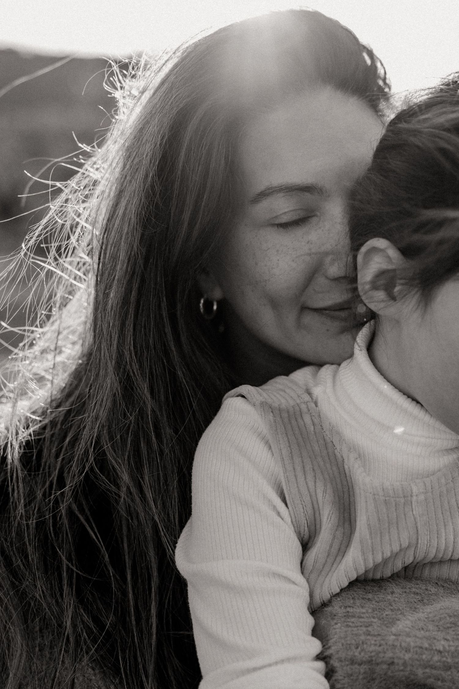 Black and white photo of a woman with freckles and long hair tenderly embracing a child in warm sunlight.