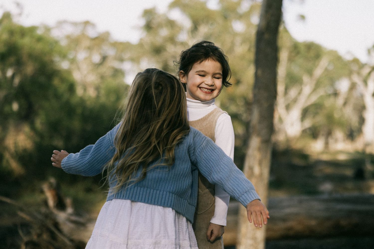 Two young girls hugging and laughing together outdoors in a sunlit forest setting at You Yangs Regional Park.