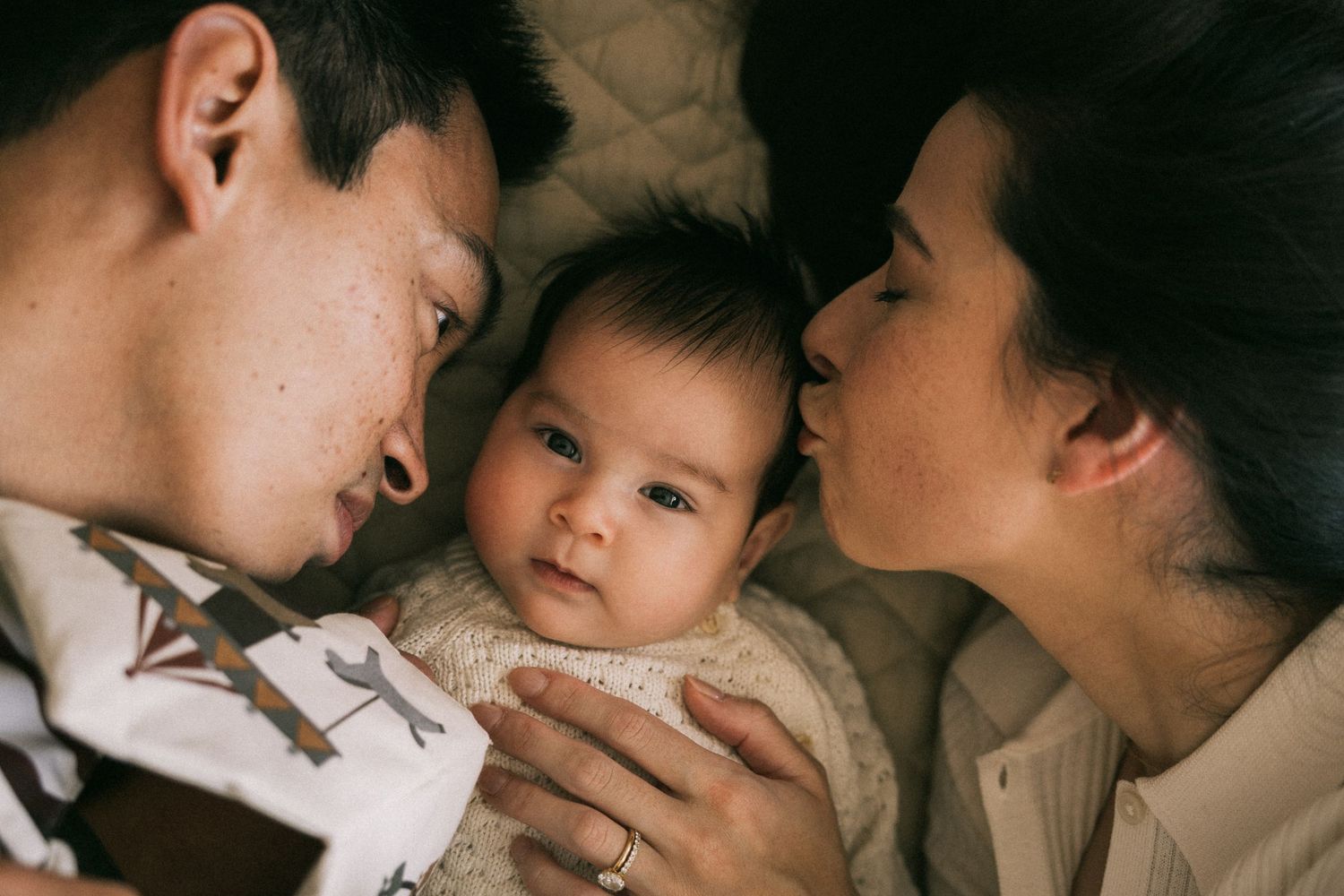 Parents lying down with their newborn baby between them, mother kissing the baby.