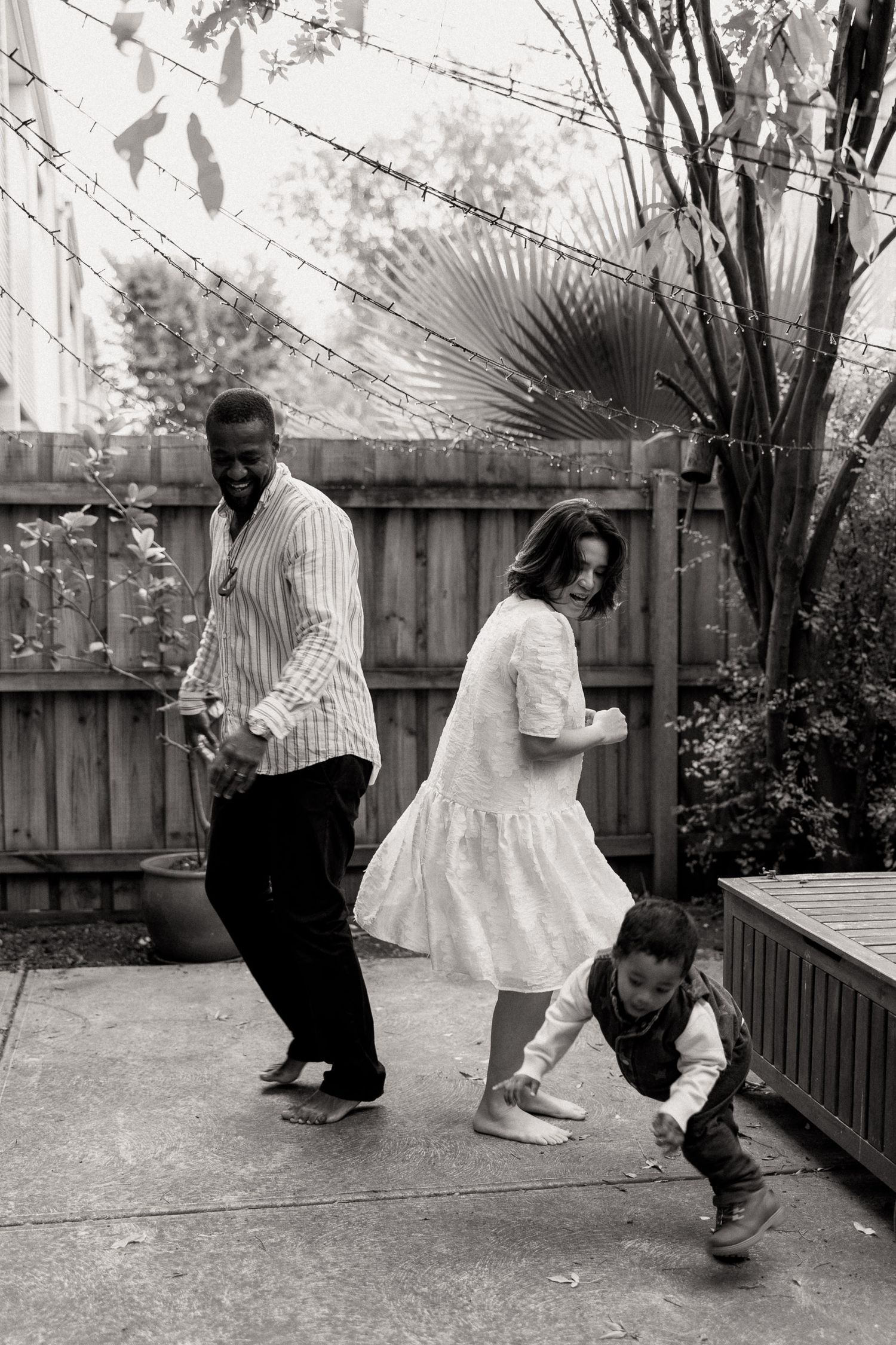 Black and white photo of a family dancing together in a backyard patio, with two adults and a young child.