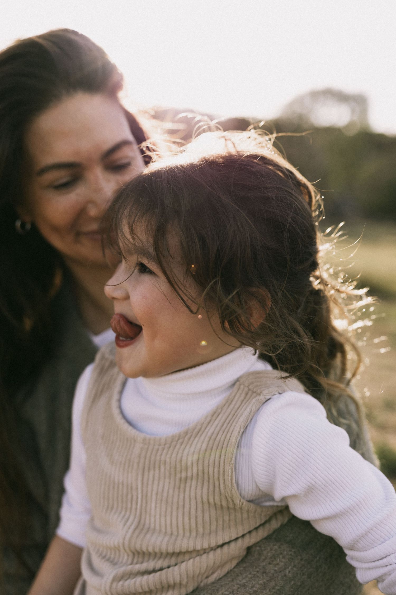 A joyful toddler girl laughs outdoors in golden sunlight while a smiling brunette woman holds her from behind at You Yangs Regional Park.