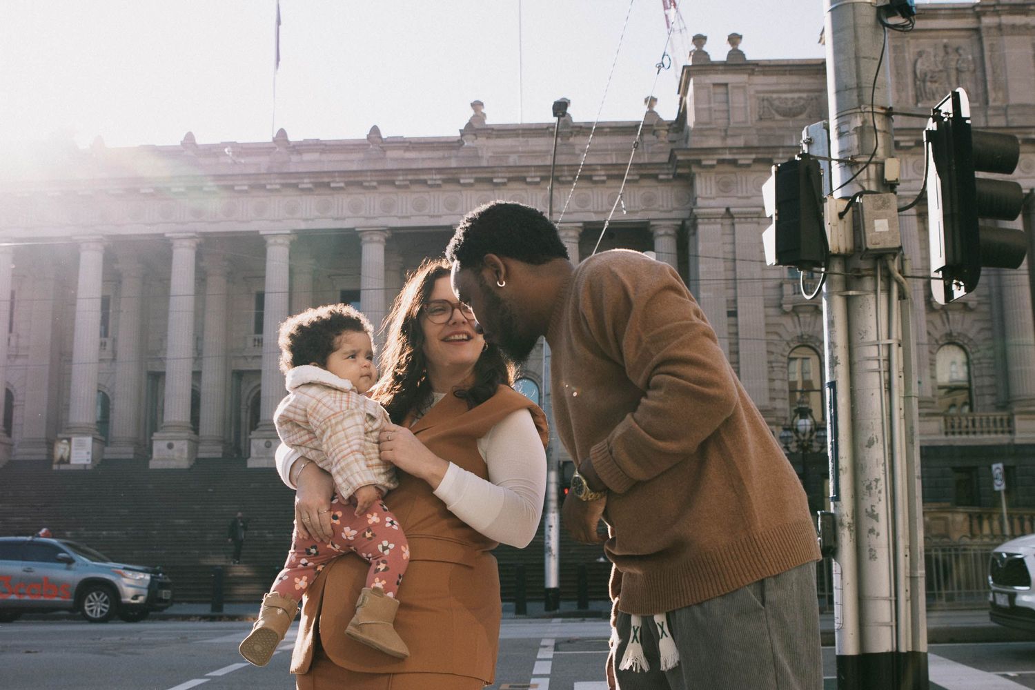 Happy family with baby girl sharing a tender moment on a sunny city street near a historic Victoria Parlament building in Melbourne CBD.