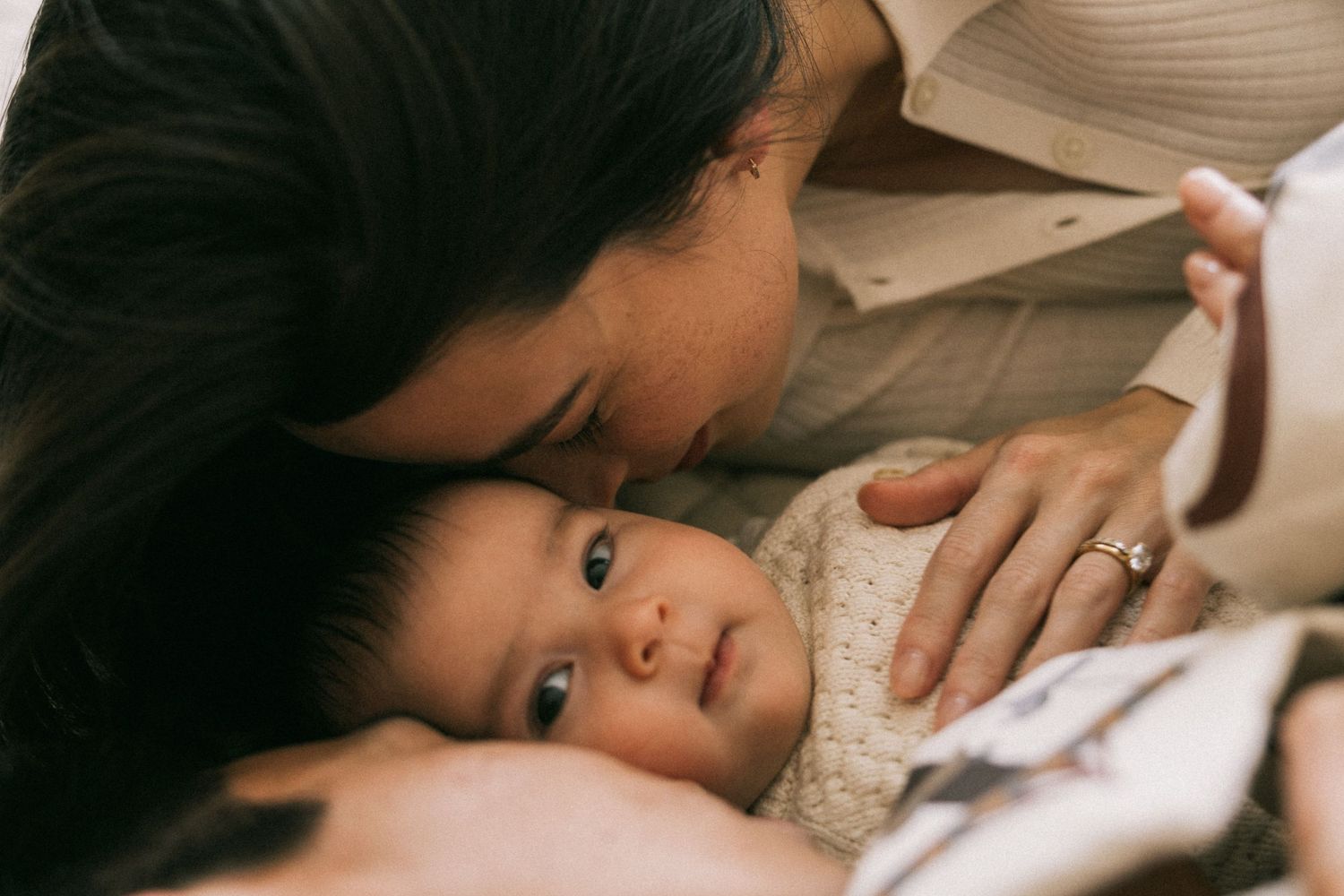 A mother tenderly kisses her newborn baby's forehead while the infant gazes upward with curious eyes in home newborn session in Altona.