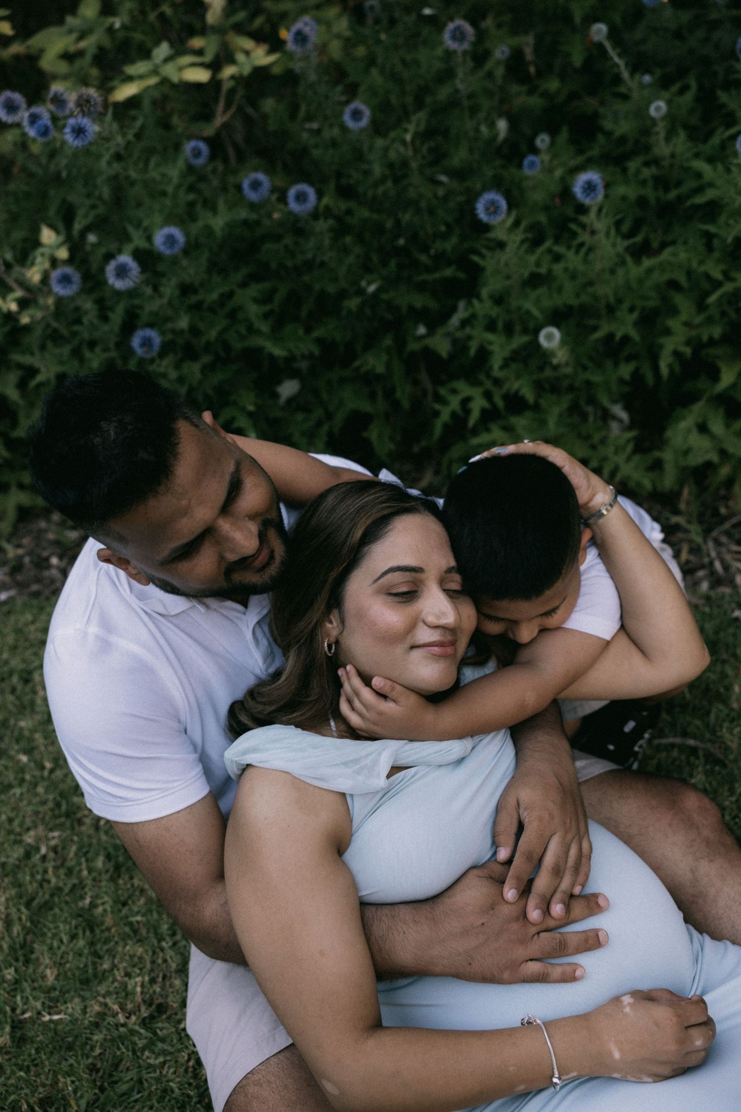 Pregnant woman sits outdoors with husband and child embracing her, surrounded by lush greenery and blue flowers in Williamstown.