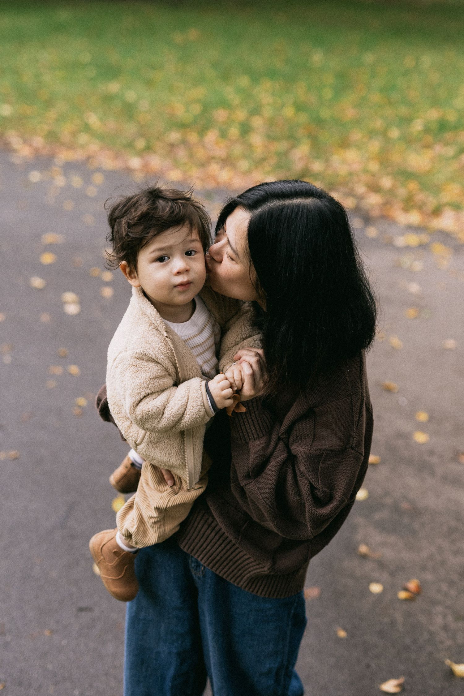 A mother kisses her toddler's cheek while holding them in Carlton Gardens during autumn.