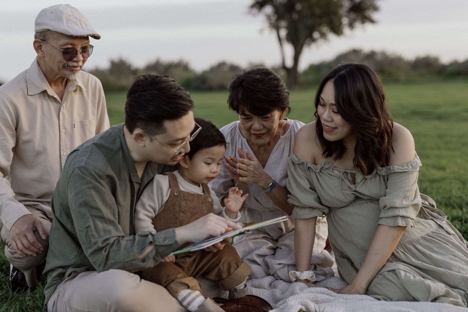 Multi-generational Asian family sitting on grass in Williamstown park, sharing a joyful moment reading a book together.