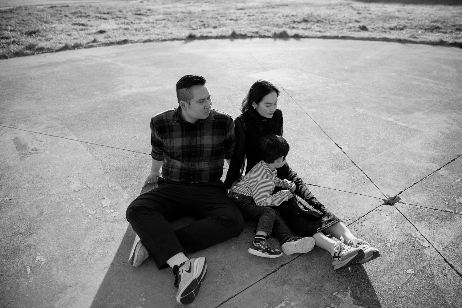 Black and white photo of a family of three sitting together on a concrete surface outdoors in Carlton.