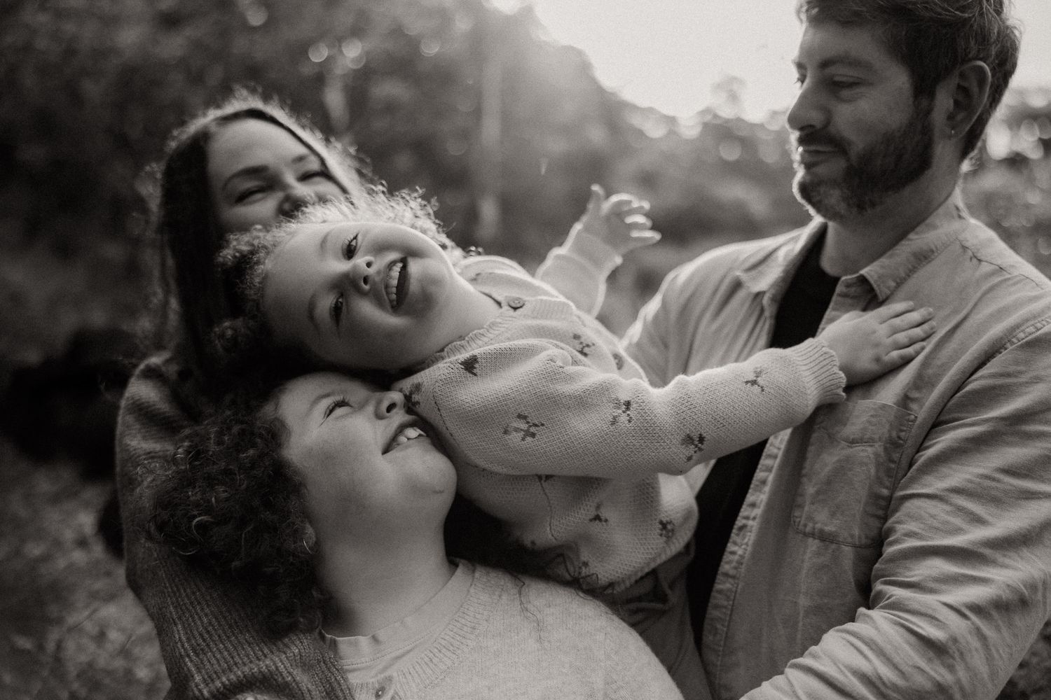 Black and white photo of a joyful family of four laughing together outdoors in Newport Lake Reserve, parents holding a giggling child.