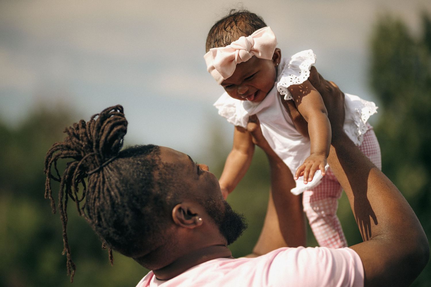 Loving father lifting smiling baby girl dressed in white outdoors on a sunny day.